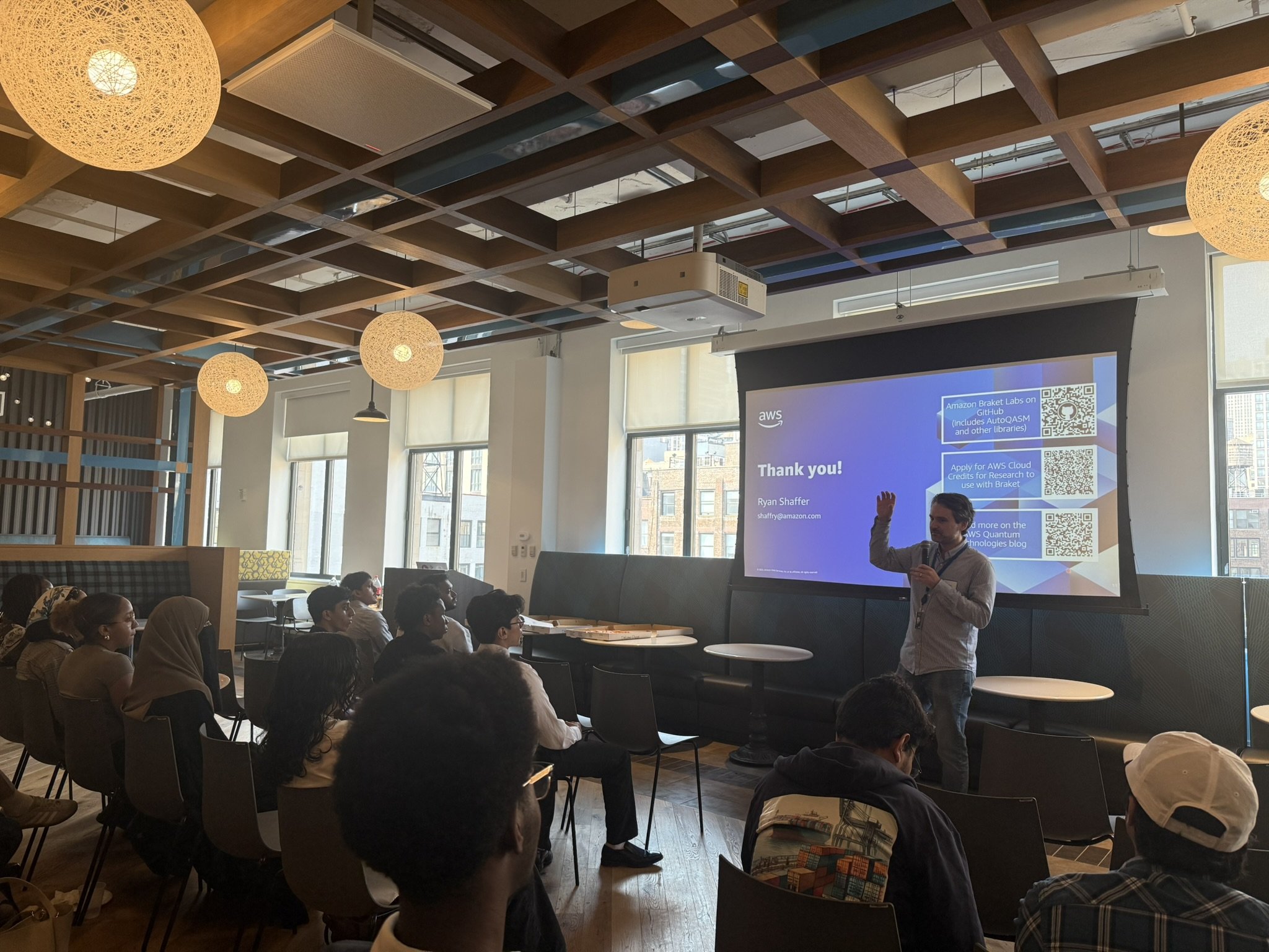 A man giving a presentation to an audience in a modern room with large windows. The screen displays a thank you slide with QR codes and contact information.