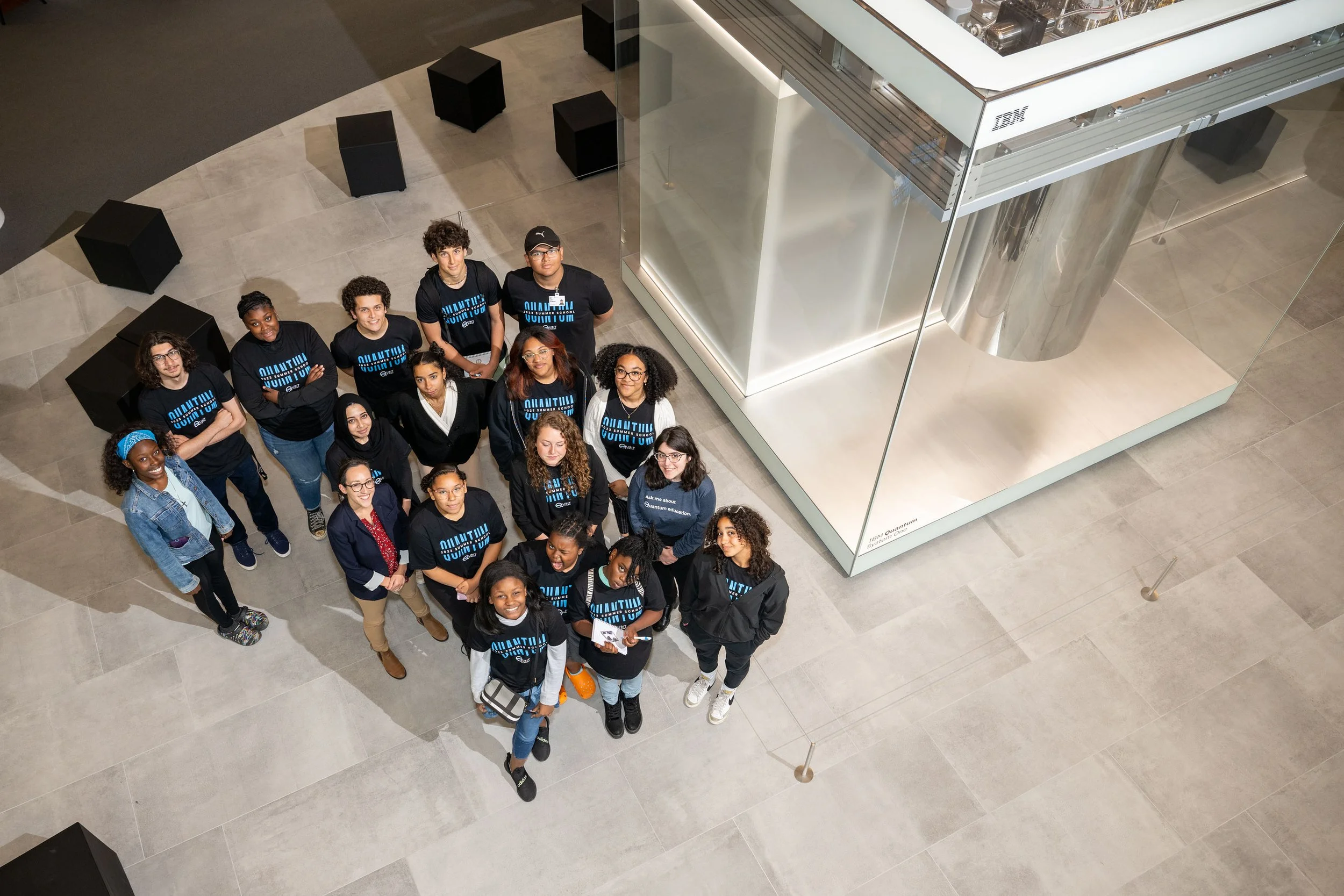 Group of diverse young people standing in a museum or science center, looking up at the camera, with a large glass exhibit case nearby.