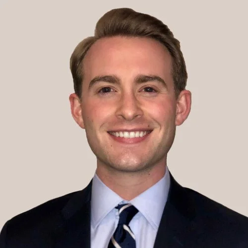Portrait of a young man with light brown hair wearing a dark suit, light blue shirt, and striped tie, smiling against a plain background.