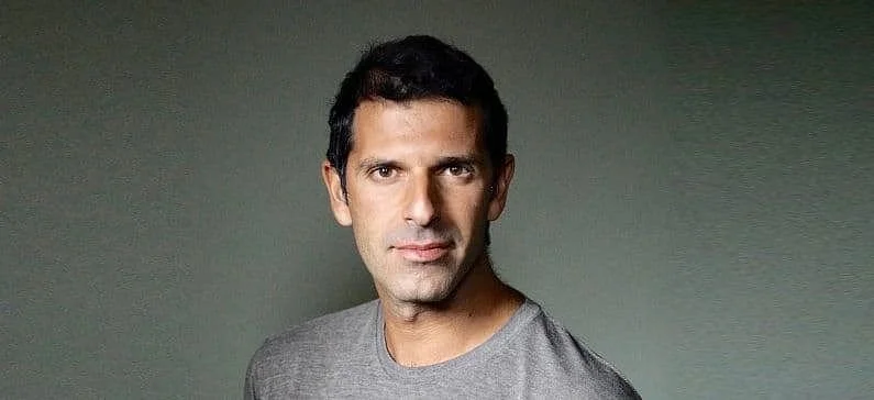 Close-up of a man with short dark hair and tan skin, wearing a gray shirt, looking directly at the camera against a plain green background.