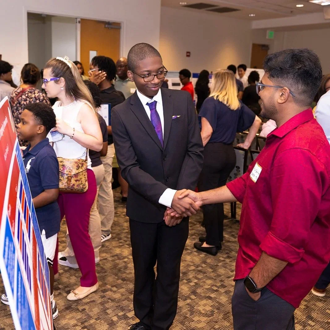 A group of diverse people at a professional event, with two men shaking hands in the foreground, one dressed in a suit and the other in a red shirt, engaging in conversation. Several others are seen mingling in the background.