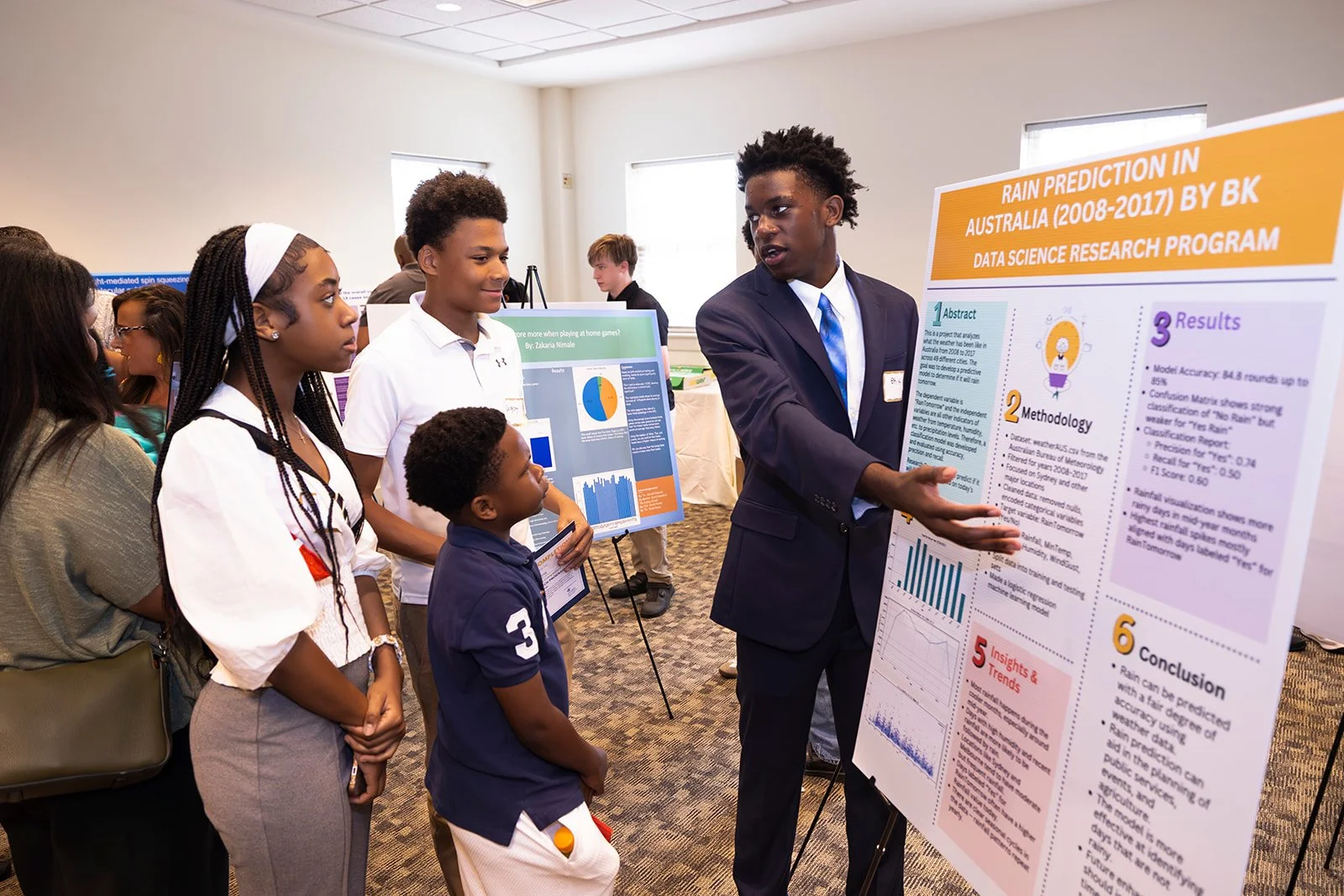 Young man in a suit presenting a research poster at a science conference, surrounded by a group of young students observing and listening.