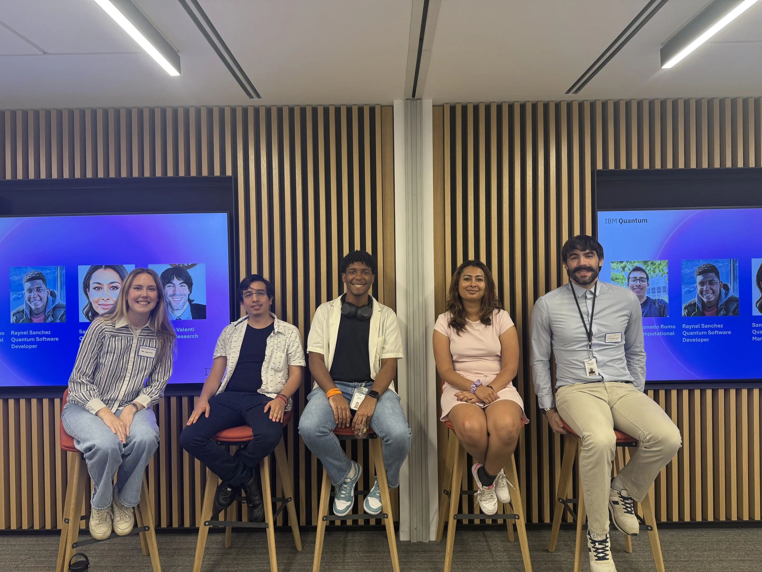 Group of five young adults seated on high stools in front of a wooden paneled wall with two large digital screens displaying profiles of individuals with names and titles.