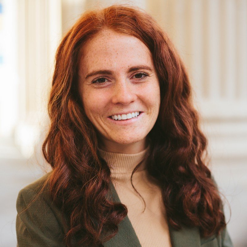 Close-up photo of a young woman with red, curly hair, freckles, and a bright smile, wearing a beige turtleneck and blazer, in a well-lit indoor setting.