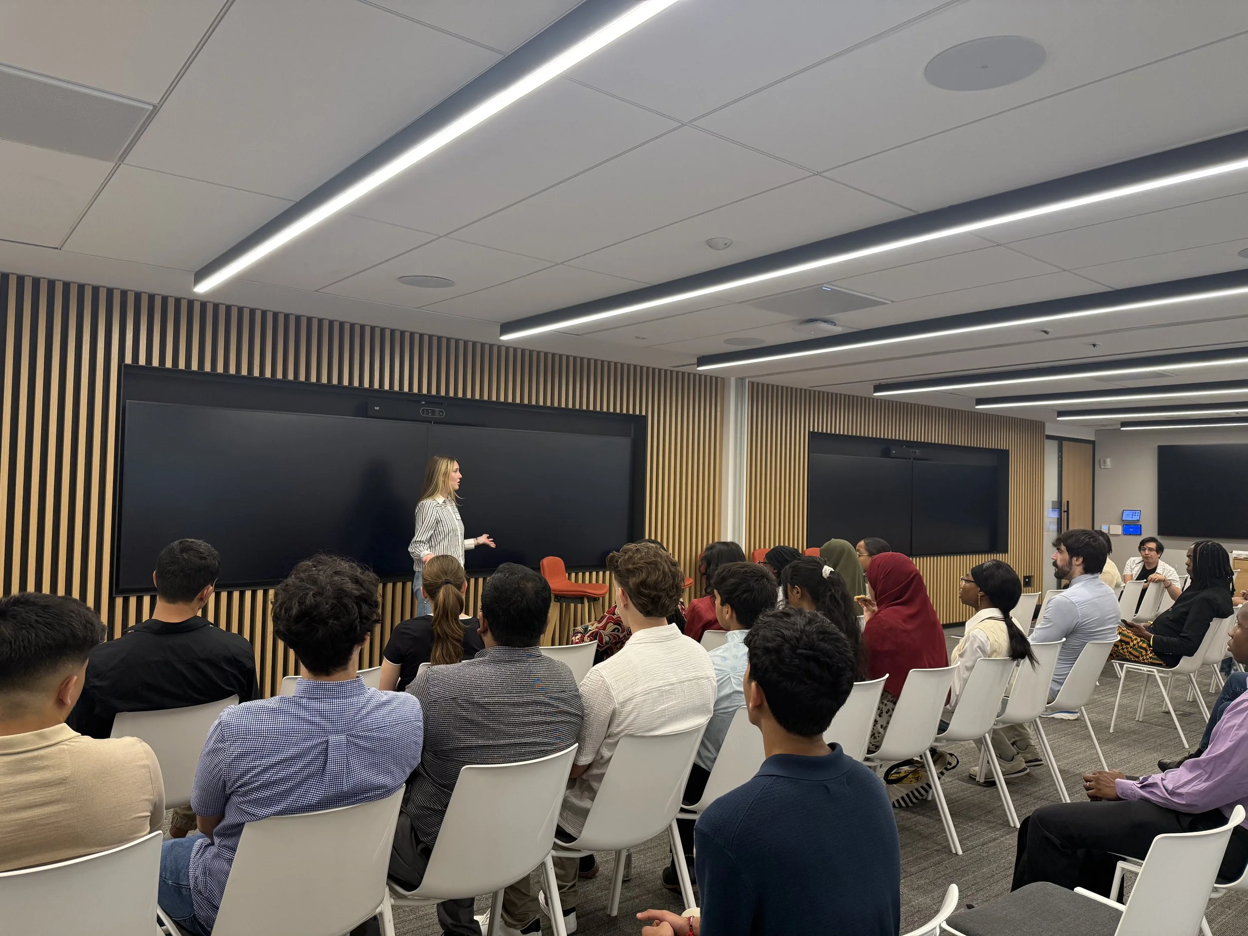 A woman is standing at the front of a room giving a presentation to a diverse audience seated in white chairs. The room has wood-paneled walls and large black screens behind her.