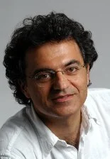A man with dark, curly hair and glasses, wearing a white shirt, smiling slightly.