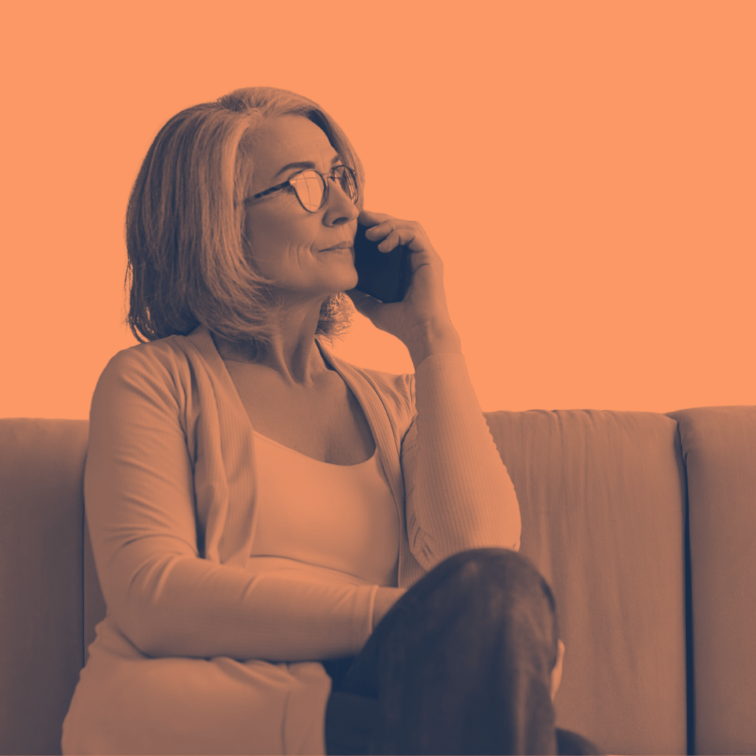 A middle-aged woman with glasses and shoulder-length hair sitting on a couch, talking on a cordless phone, smiling, wearing a light-colored cardigan and top.