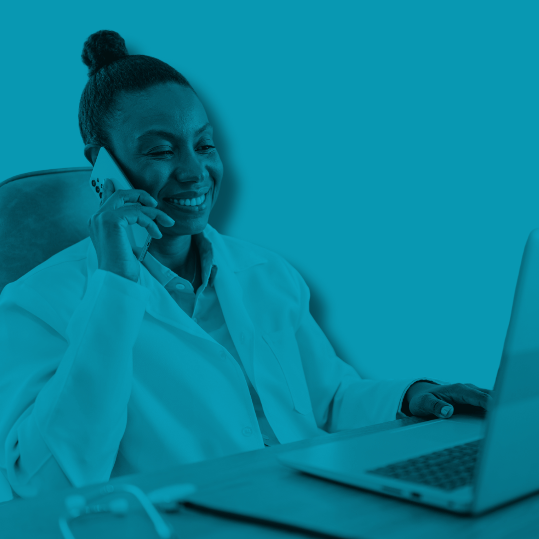 A woman with a bun hairstyle smiling and talking on a mobile phone while sitting at a desk with a laptop.