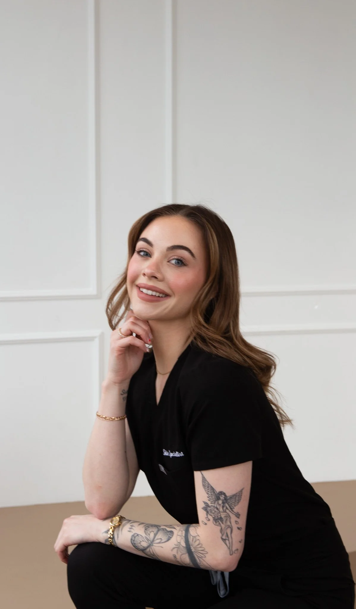 A young woman with brown hair in loose waves, wearing a black T-shirt, kneeling with her right arm resting on her knee and her chin on her hand, smiling at the camera.