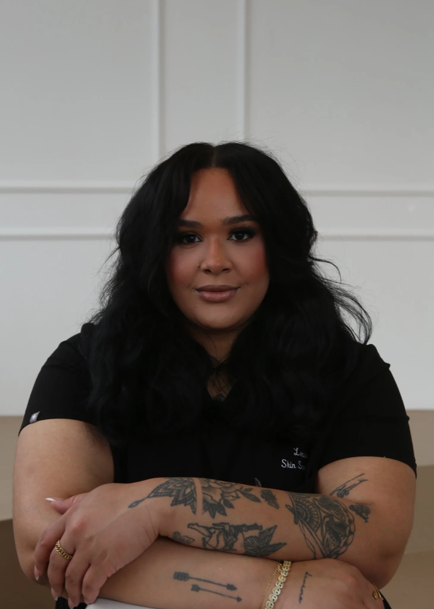 A woman with black wavy hair and tattoos on her arms, sitting with arms crossed, wearing a black top, in front of a white wall.