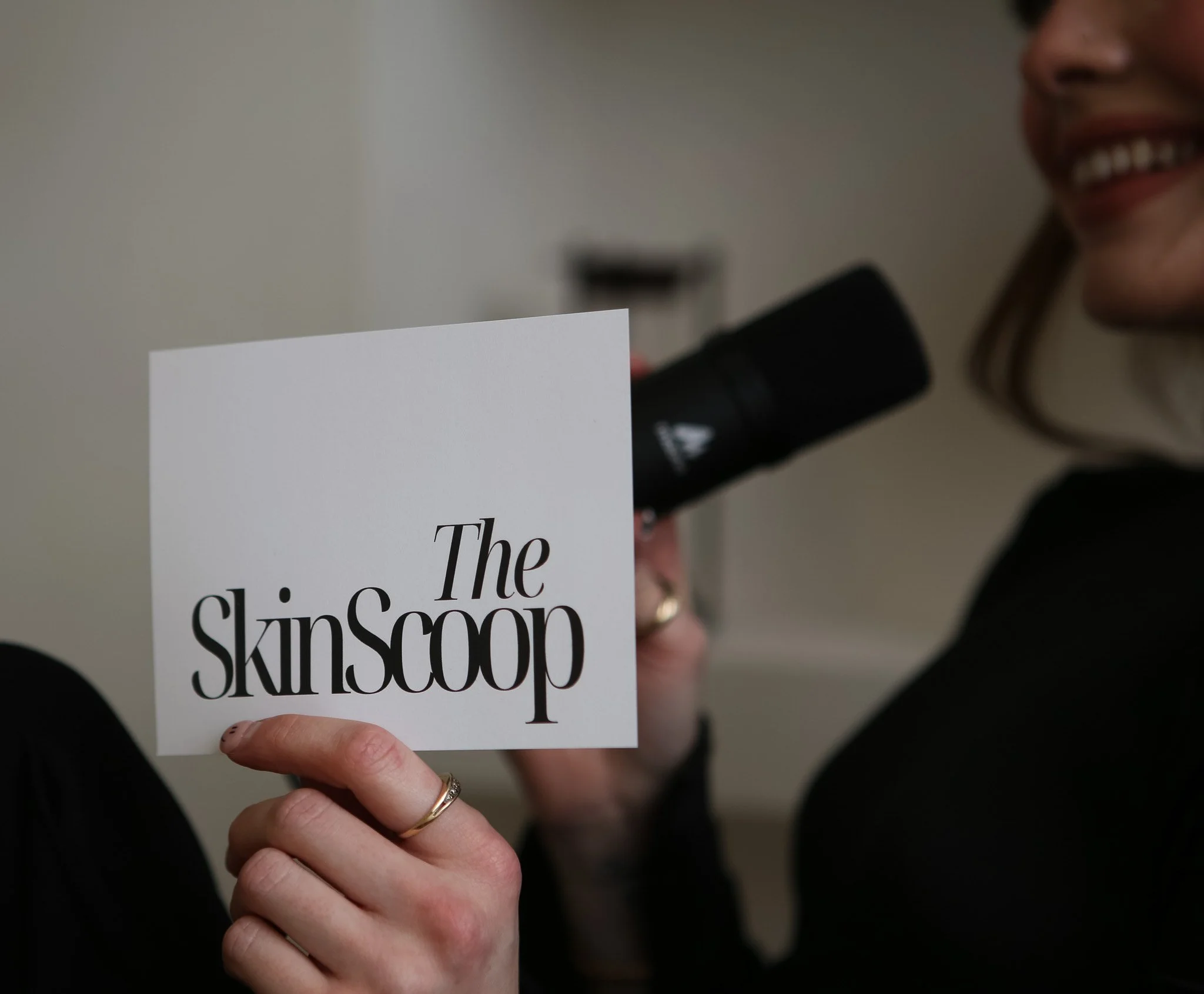 A woman smiling and holding a sign that reads "The SkinScoop" in front of a microphone.
