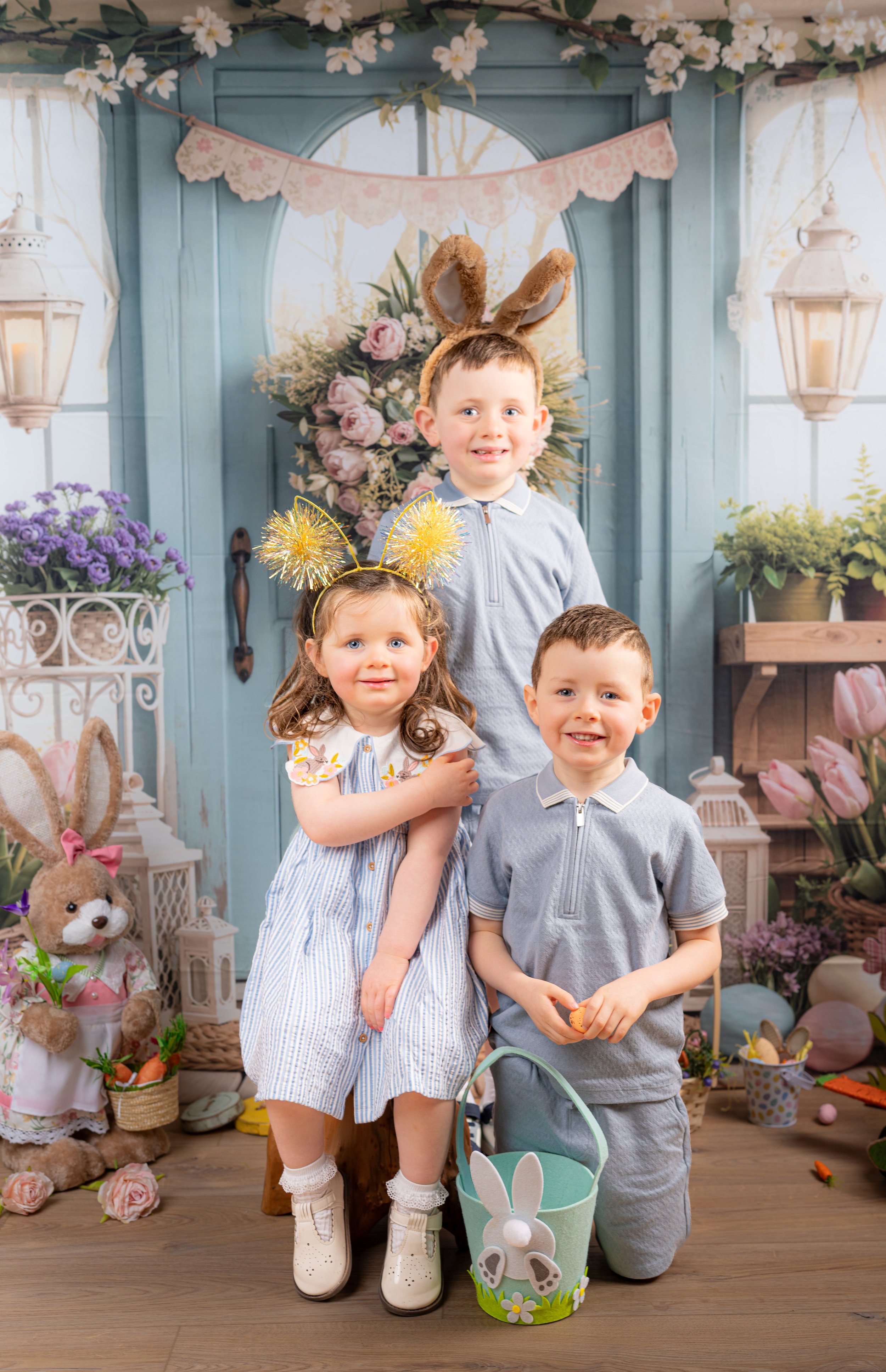 Three children celebrating Easter in a decorated room with bunny ears, flowers, and Easter-themed decorations. One girl wearing glittery bunny ears and a dress, a boy kneeling with a basket, and another boy standing with bunny ears on his head. They are smiling and surrounded by pastel-colored decorations, including flowers, lanterns, and plush bunny toys.