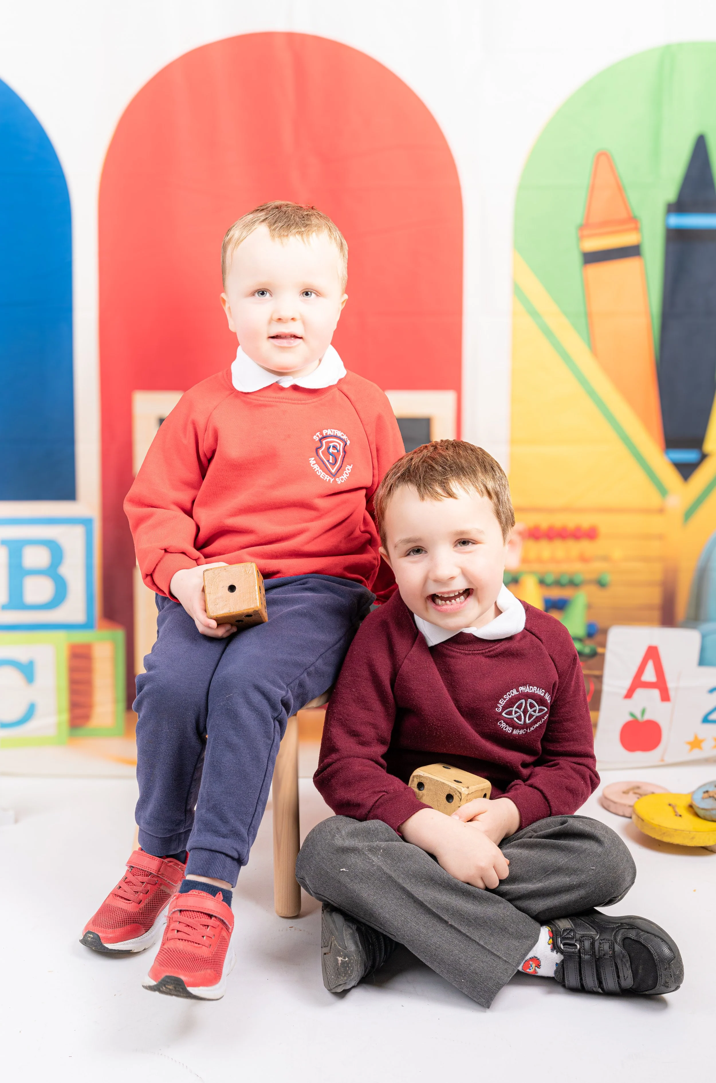 Two young boys in school uniform sitting in front of colorful classroom background with alphabet blocks and toys, holding wooden dice, one boy sitting on the floor and the other on a small stool.
