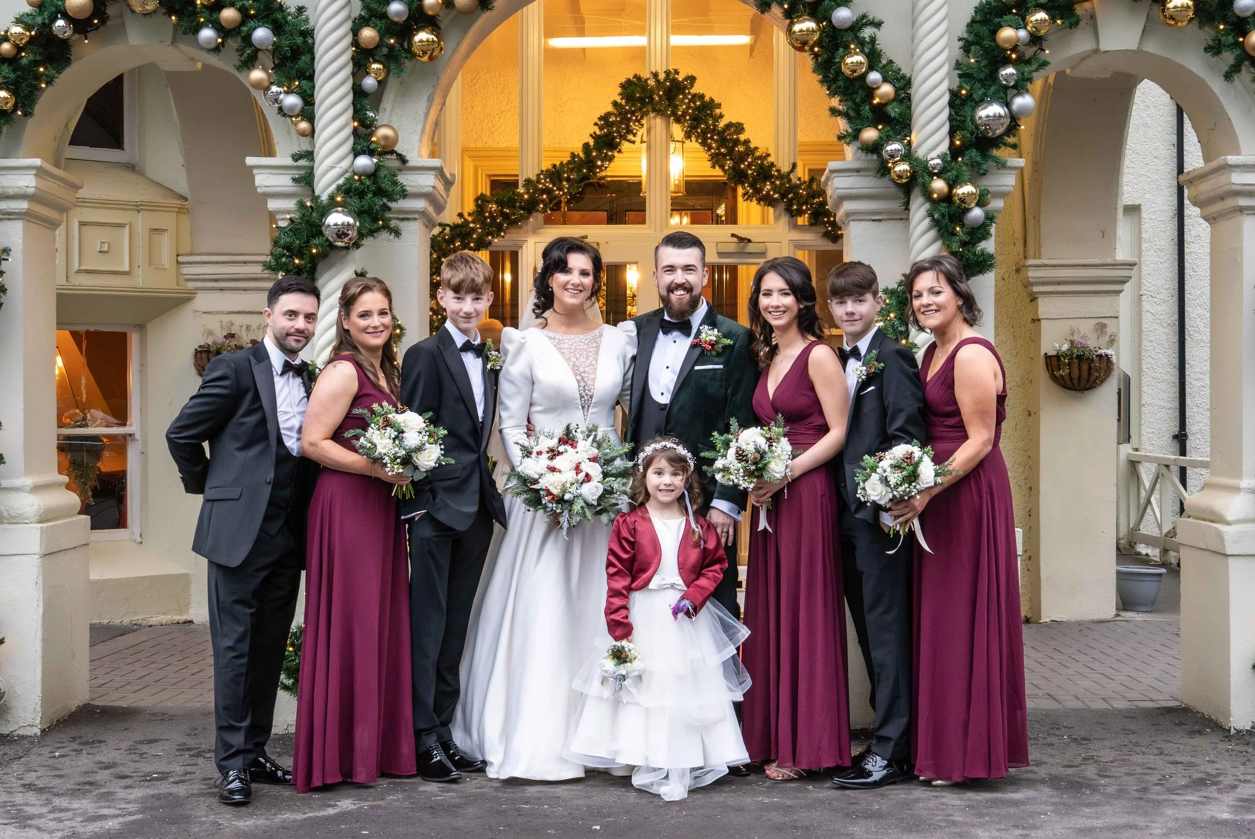 A wedding party standing outside a decorated building with Christmas garlands and ornaments, including brides, grooms, children, and bridesmaids, all smiling and holding bouquets.