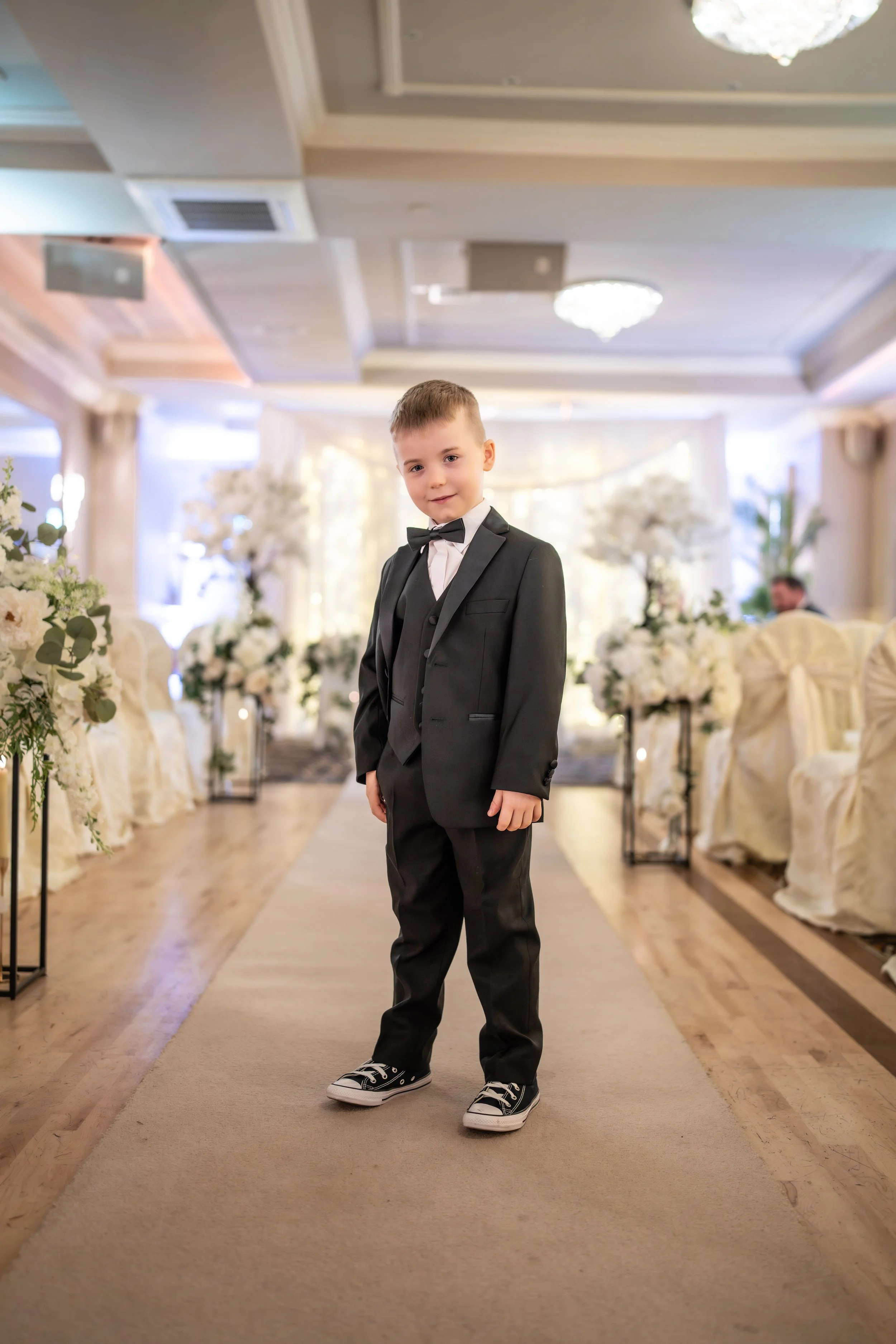 Young boy in black tuxedo with bow tie and sneakers standing on a decorated aisle at a wedding ceremony.