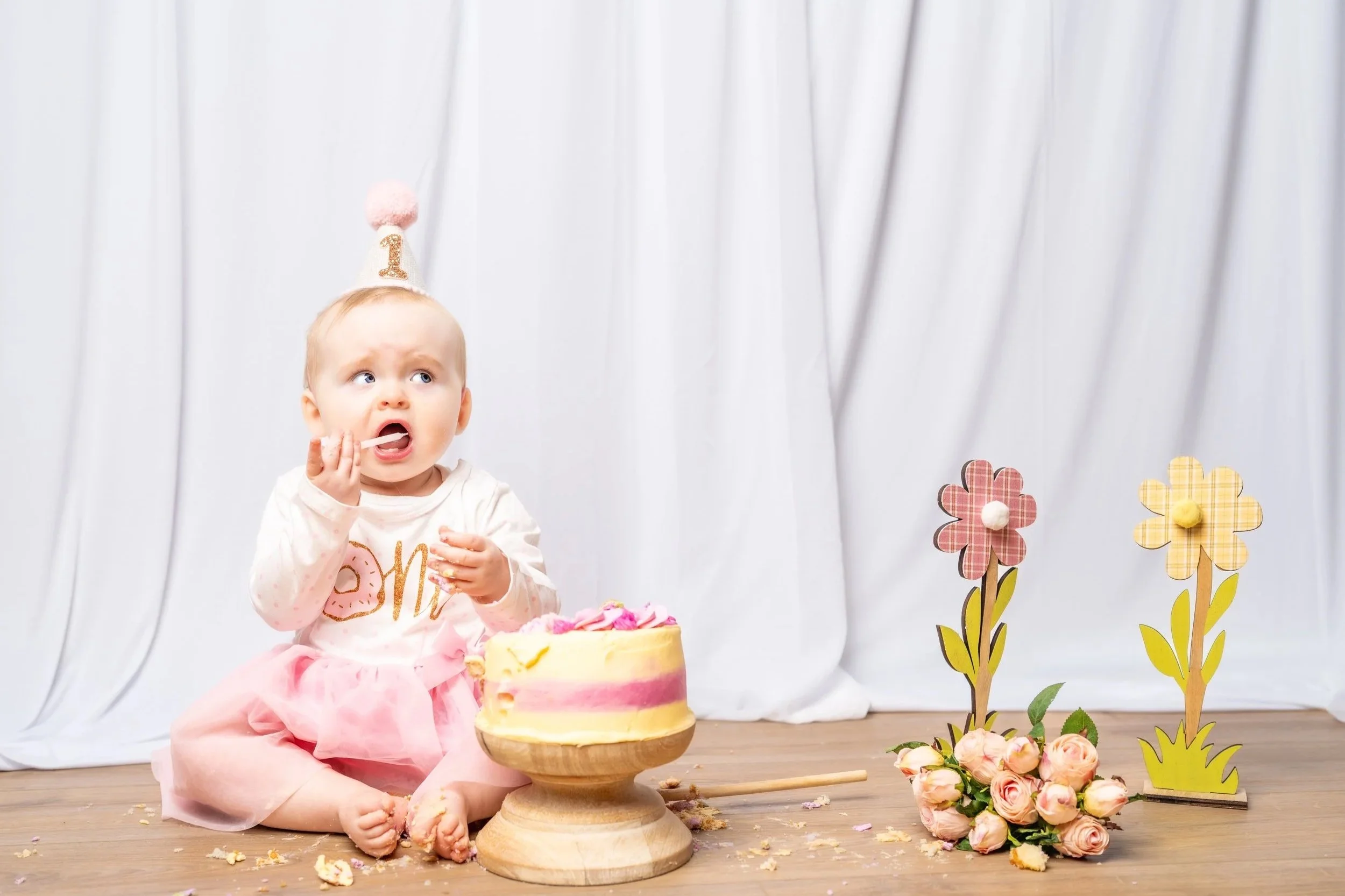 Baby girl celebrating her first birthday with a pink and yellow cake, sitting on the floor with a pink tutu, wearing a white shirt and a birthday hat with the number one, surrounded by flowers and decorative flowers on a wooden floor with a light background.