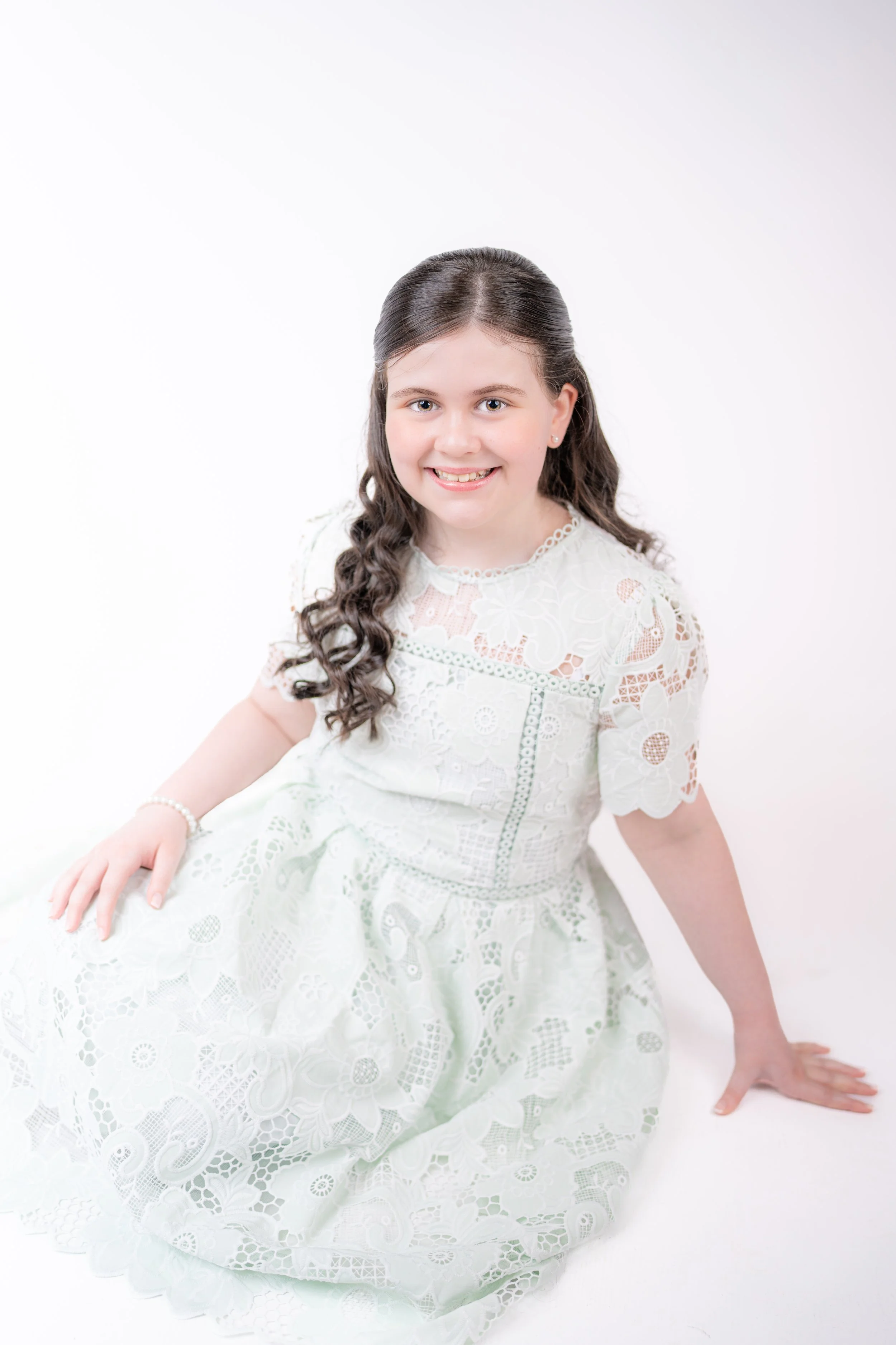 A young girl in a white dress standing on a wooden staircase, smiling, with her right hand resting on the staircase railing.