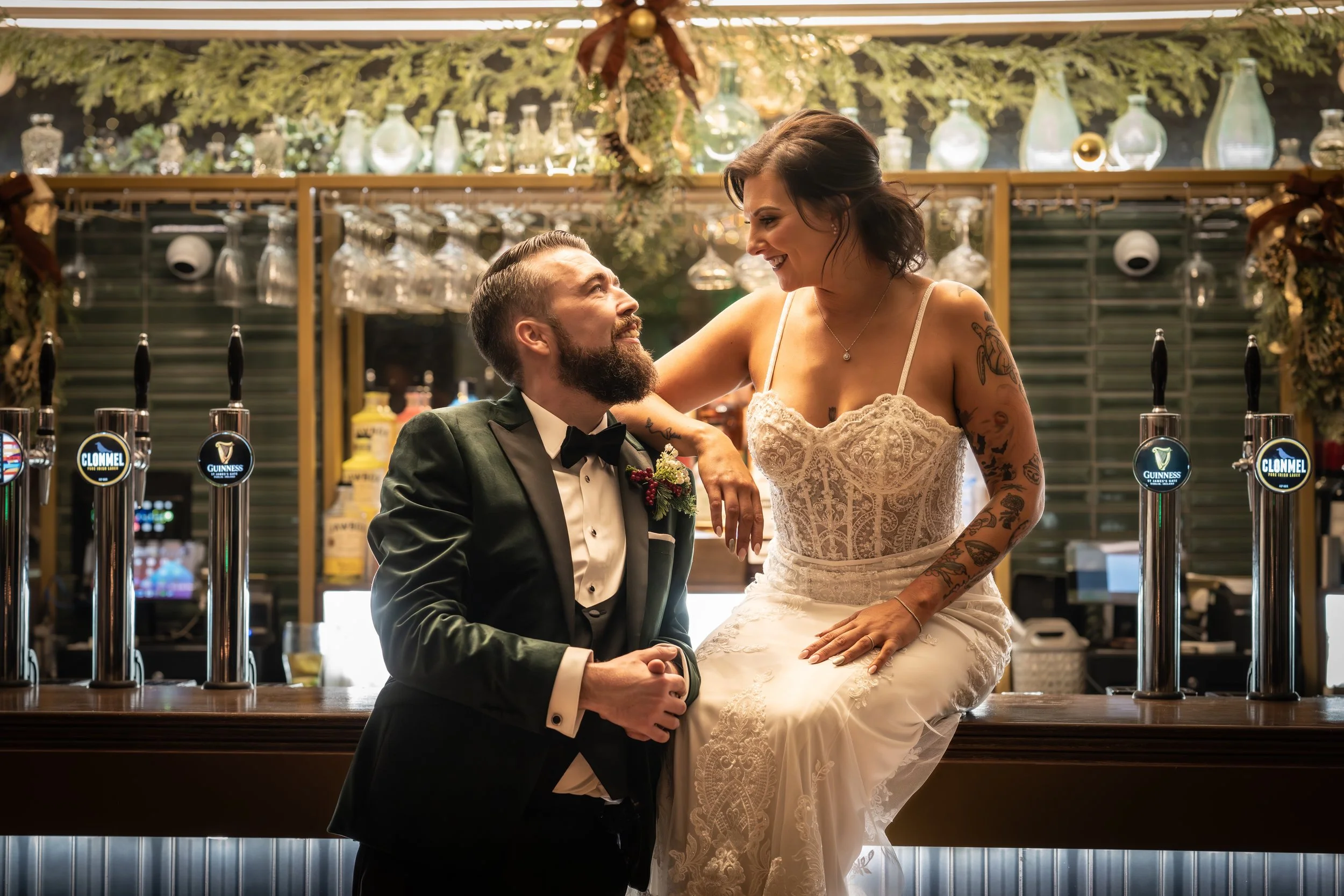 A man in a tuxedo with a beard and a woman in a wedding dress with tattoos sitting on a bar counter, smiling and looking at each other in a decorated bar setting.