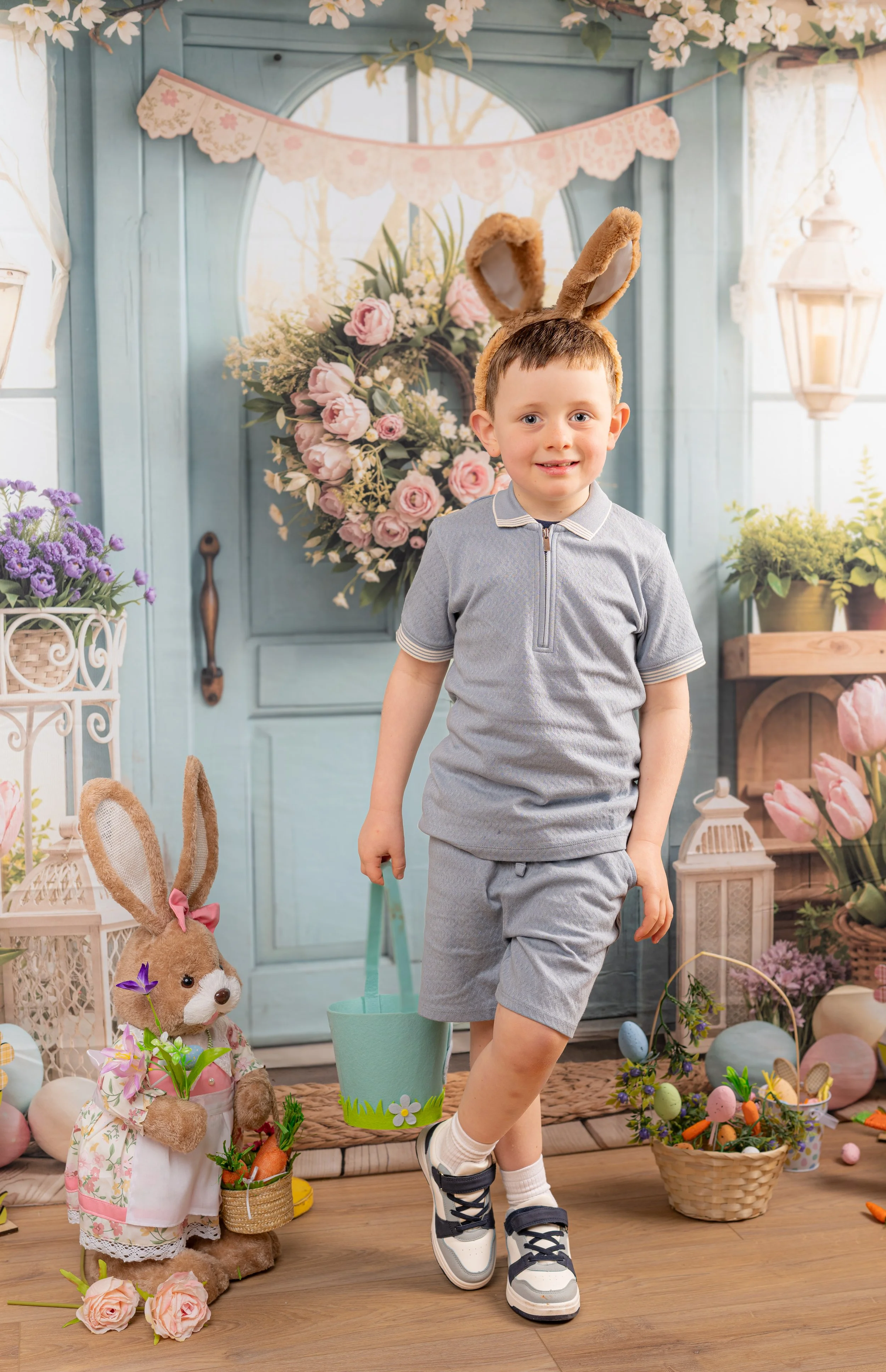 A young boy dressed casually with a bunny ear headband standing in front of a decorated Easter-themed backdrop holding a small blue bucket. The background features pastel-colored flowers, Easter eggs, a bunny plush toy with a basket of carrots, and spring-themed decorations.