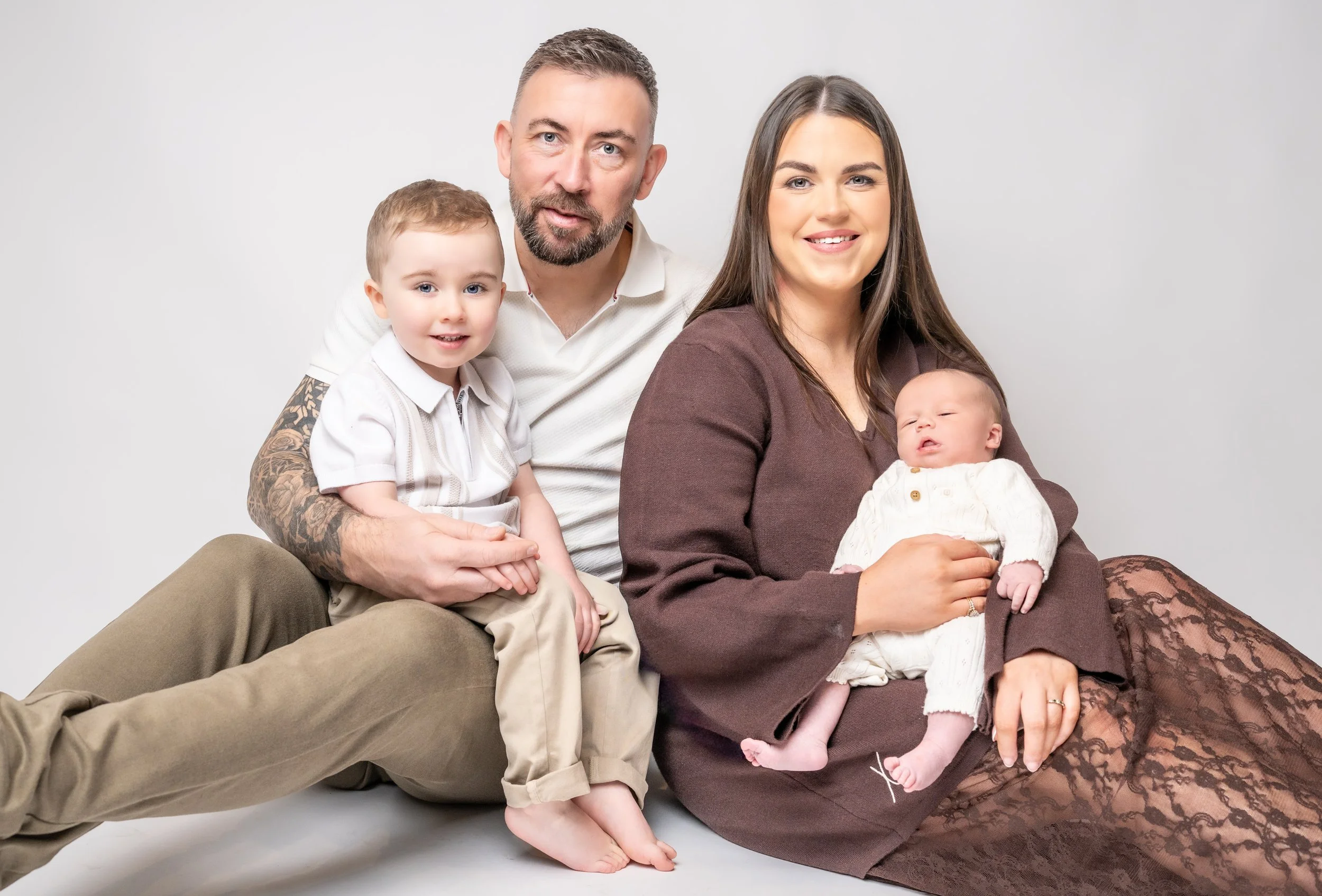 A family of four with a father, mother, and two young children, sitting against a plain light gray background.
