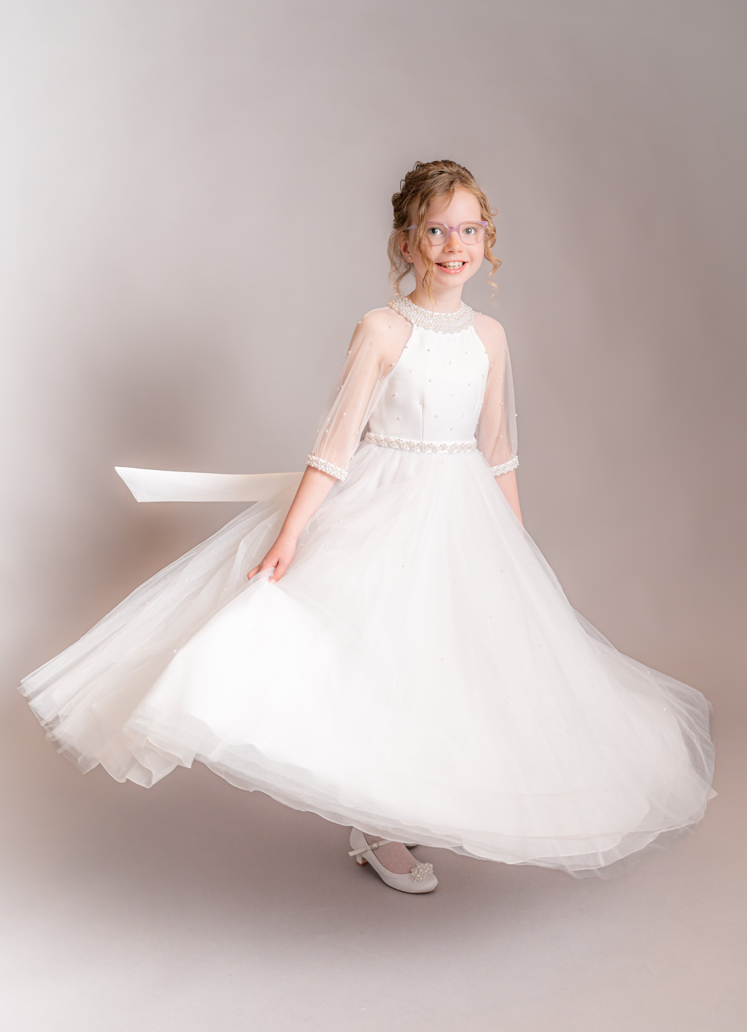 A young girl in a white dress with sheer sleeves, glasses, and a pearl necklace, smiling and posing on a light gray background.
