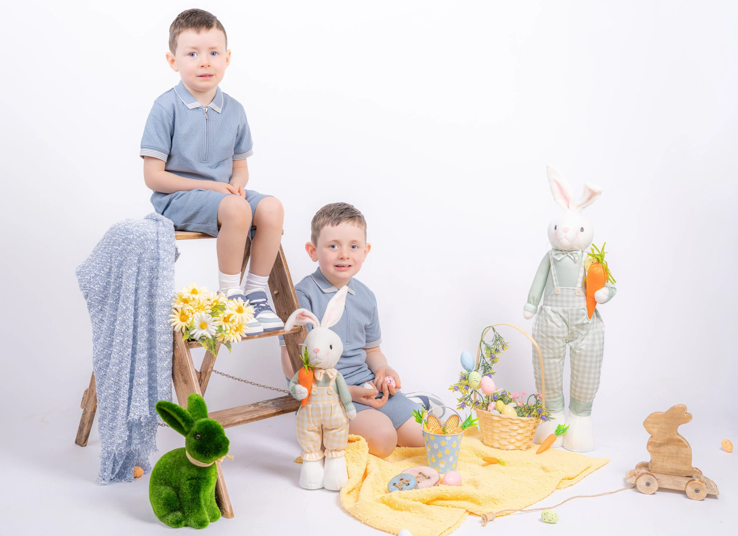 Two young boys celebrating Easter with stuffed bunny toys, decorated eggs, and spring decor on a yellow blanket, in front of a white background.
