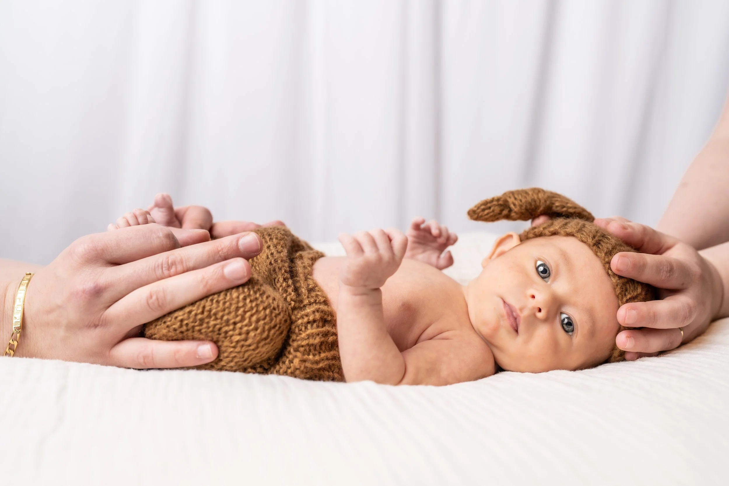 Baby lying on a white surface, wearing a brown knitted outfit and hat with ears, with adult hands gently holding and supporting the baby.