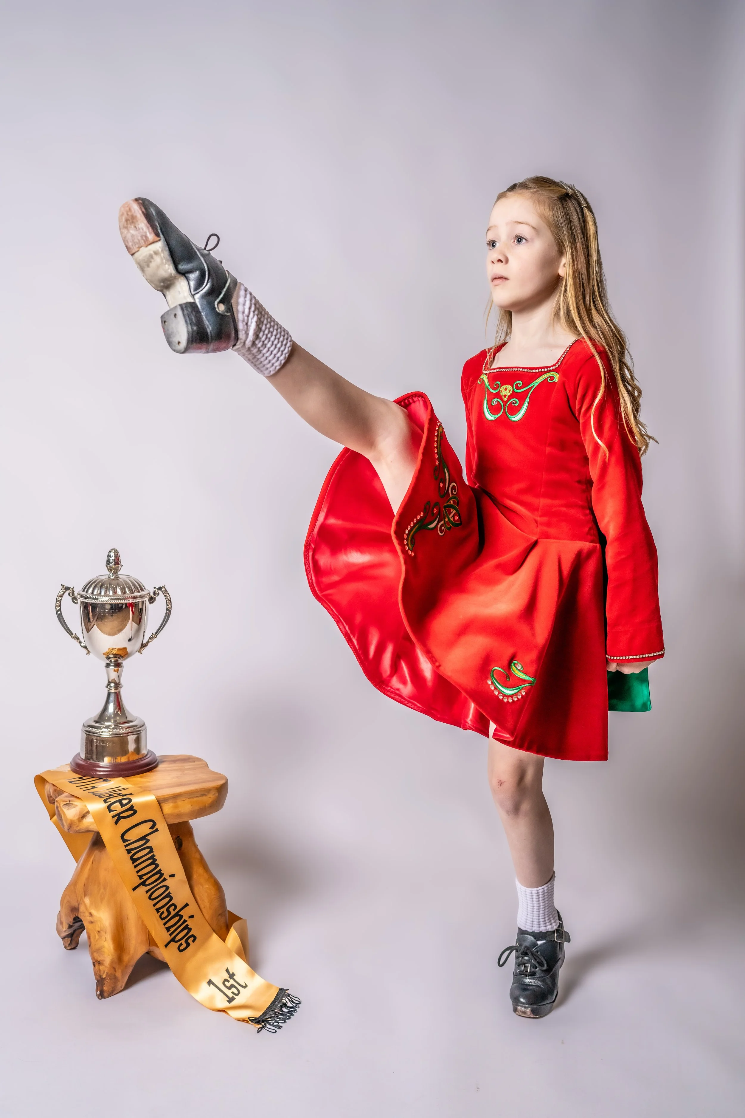 Young girl in a red dress performing a figure skating move with her leg raised high, next to a trophy on a wooden stand with a gold ribbon that reads "1st".