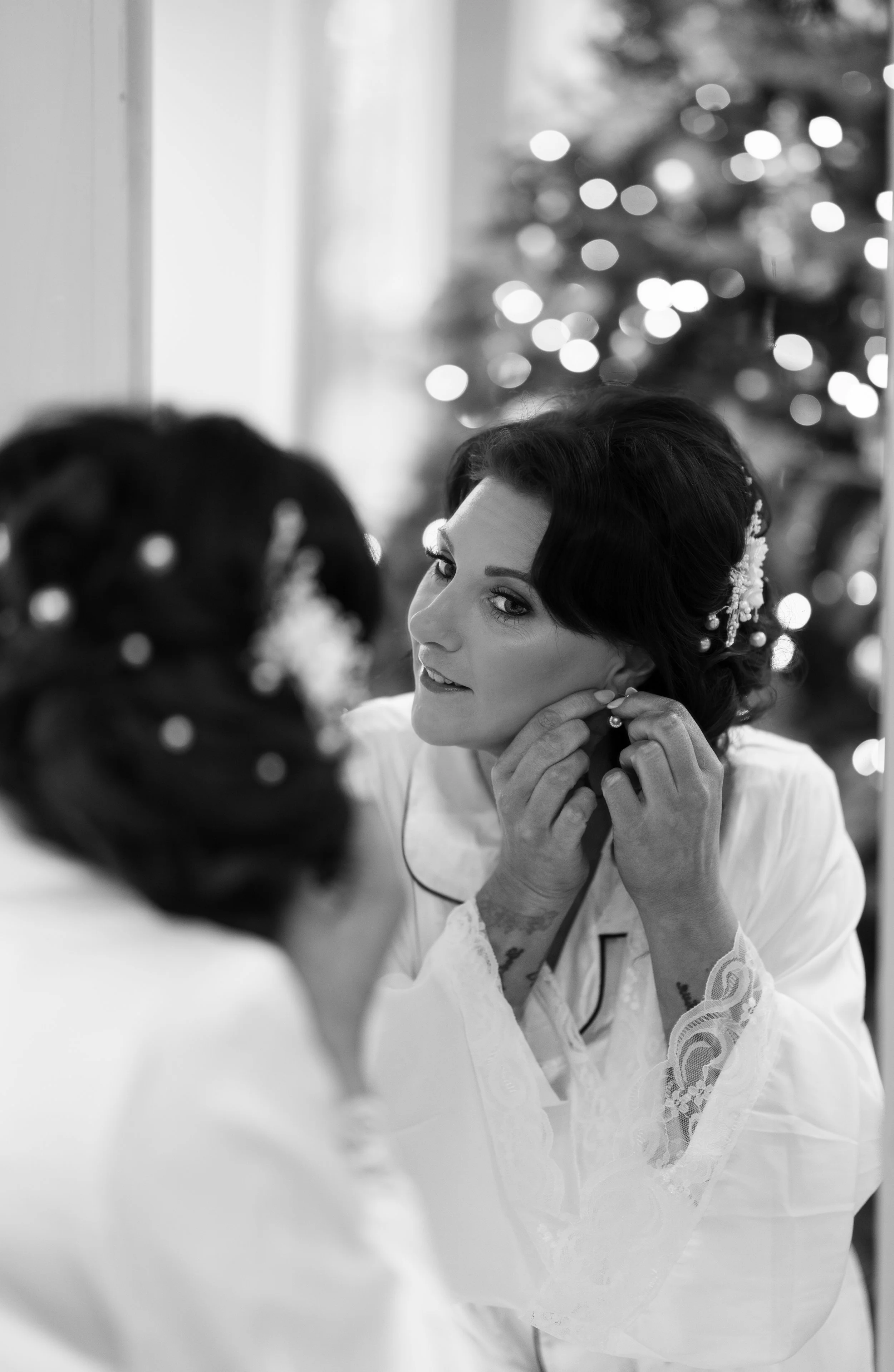 A woman with dark, wavy hair is putting on earrings while looking at her reflection in a mirror. She is wearing a light-colored, lace-trimmed robe, and a Christmas tree with lights is visible in the background.