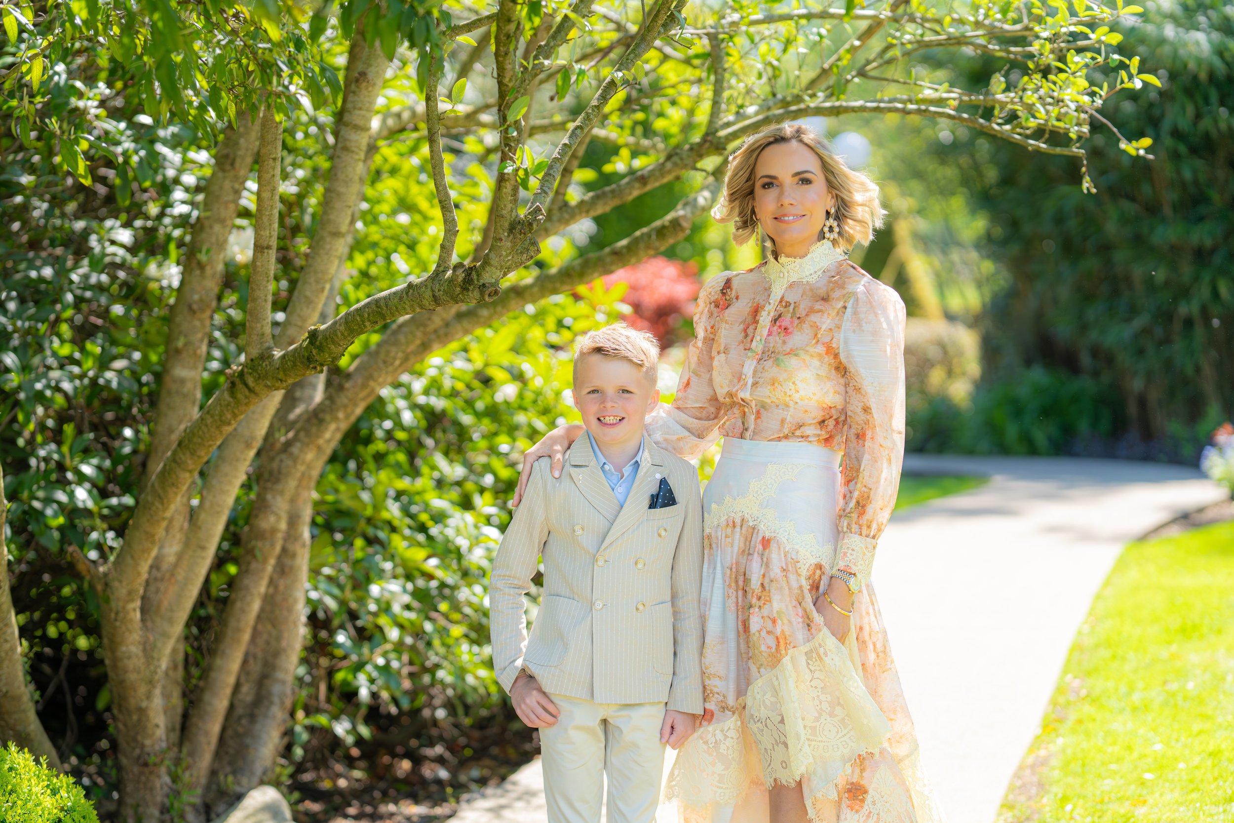 A woman and a young boy standing outdoors near a tree on a sunny day, smiling. The woman is wearing a floral dress and earrings, and the boy is in a beige pinstripe suit with a blue shirt. There is greenery in the background with sunlight filtering through the leaves.
