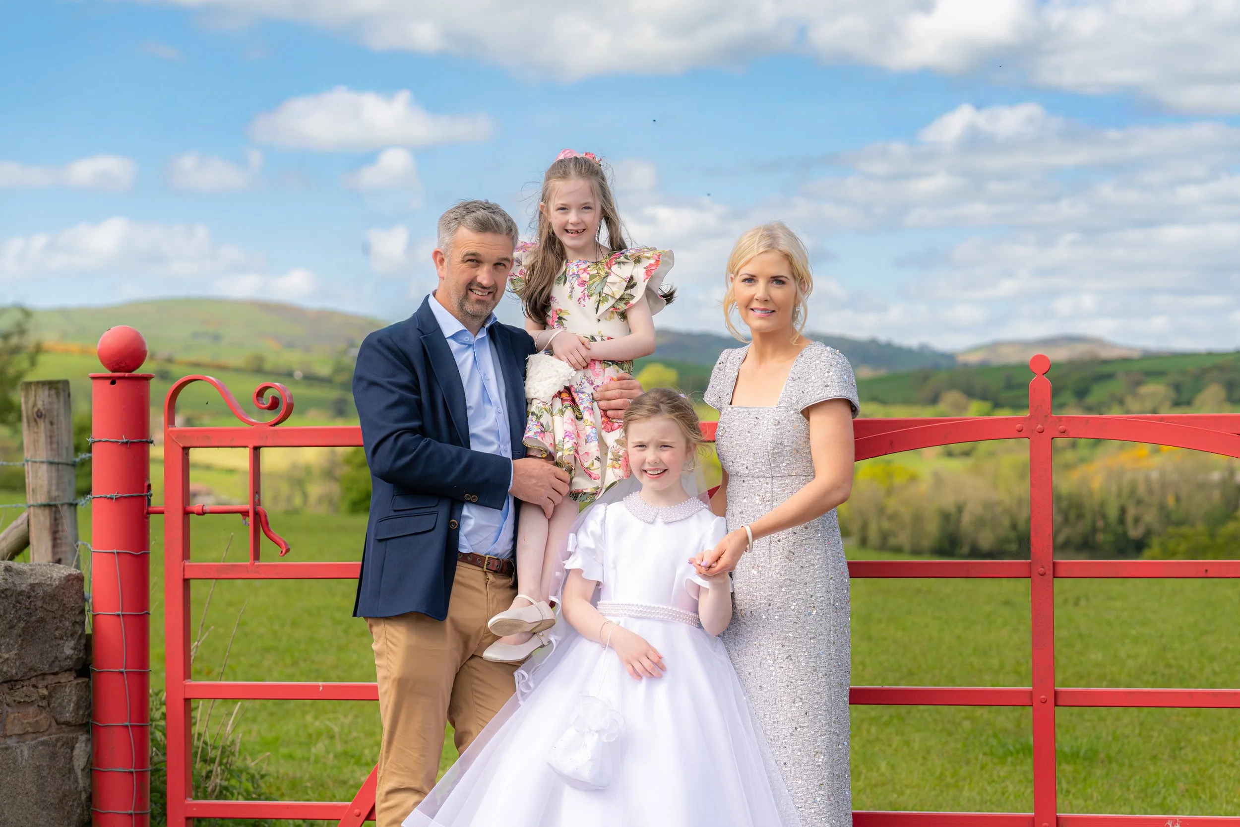 A family of five standing outdoors near a red fence, with a field and rolling hills in the background. The family includes a man in a navy blazer, a woman in a silver dress, a teenage girl in a floral dress, and two young girls in white dresses. The sky is partly cloudy.