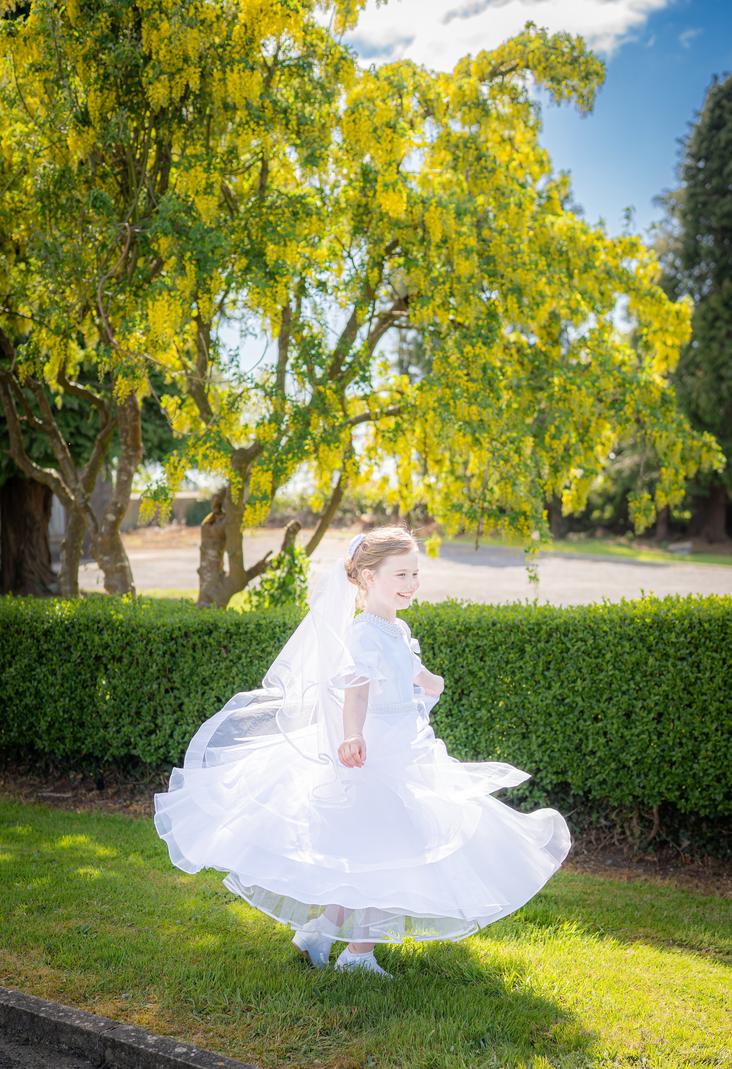 A young girl in a white dress twirling outdoors with trees and bushes in the background.