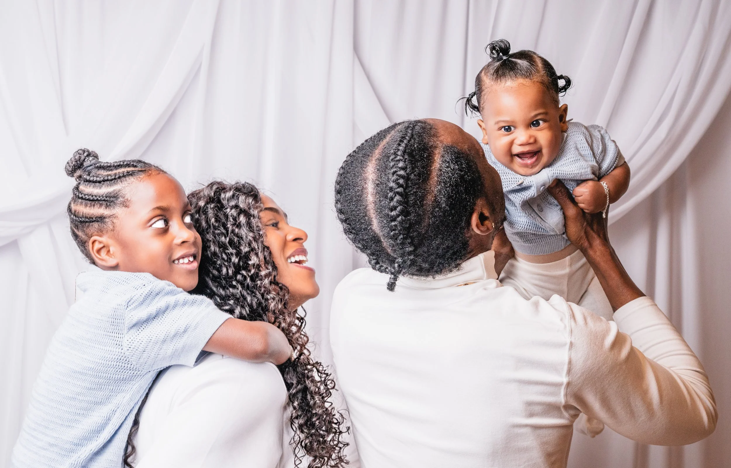 A joyful moment with three women and a young child, two women are holding the child in a loving manner, against a white curtain background.