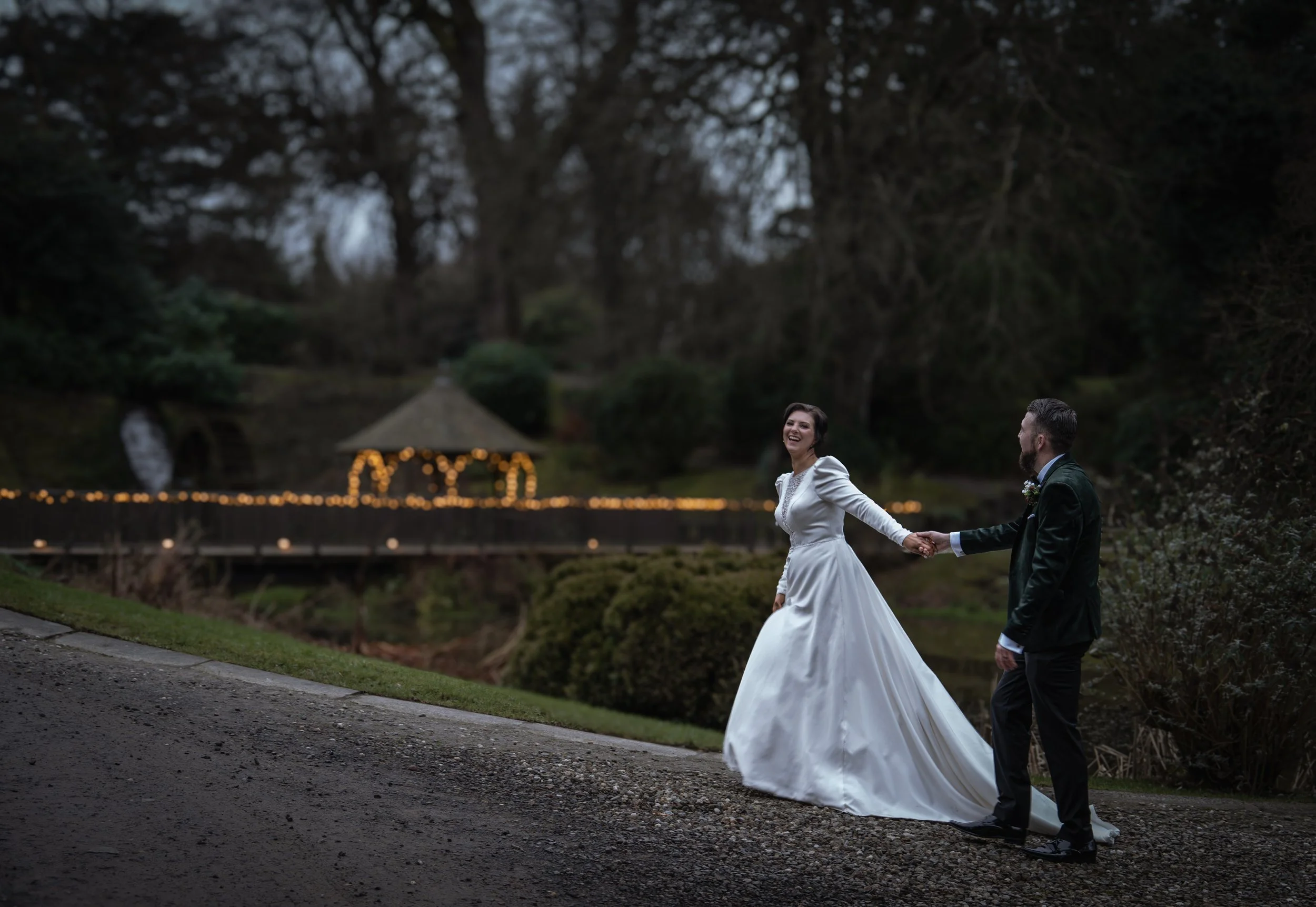 A bride and groom holding hands, walking outdoors during dusk or evening. The bride is wearing a white wedding dress and the groom a dark suit. In the background, a gazebo with string lights is visible, surrounded by trees and shrubs.