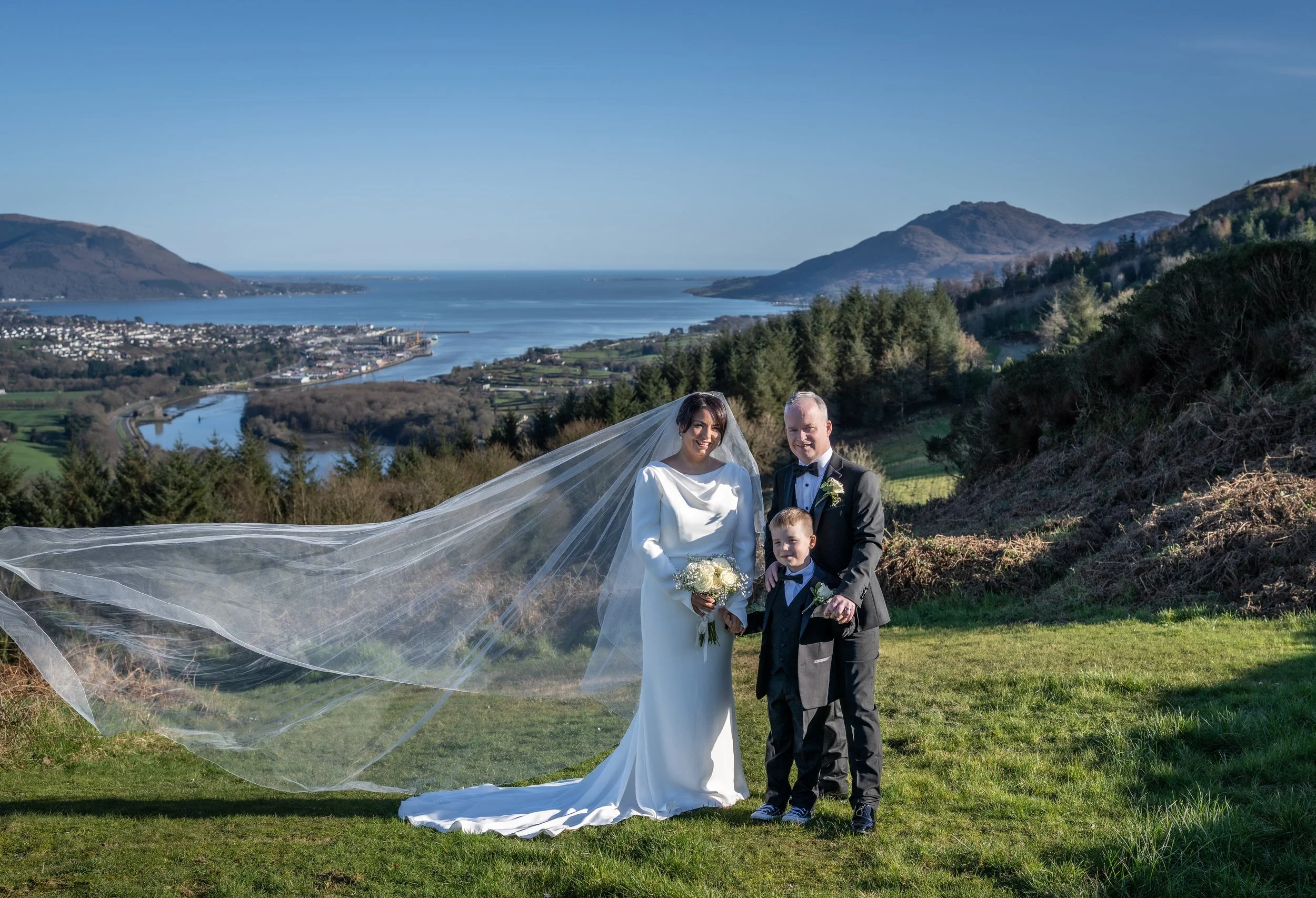 Wedding couple and young boy standing on grassy hillside with scenic lake, mountains, and trees in background.