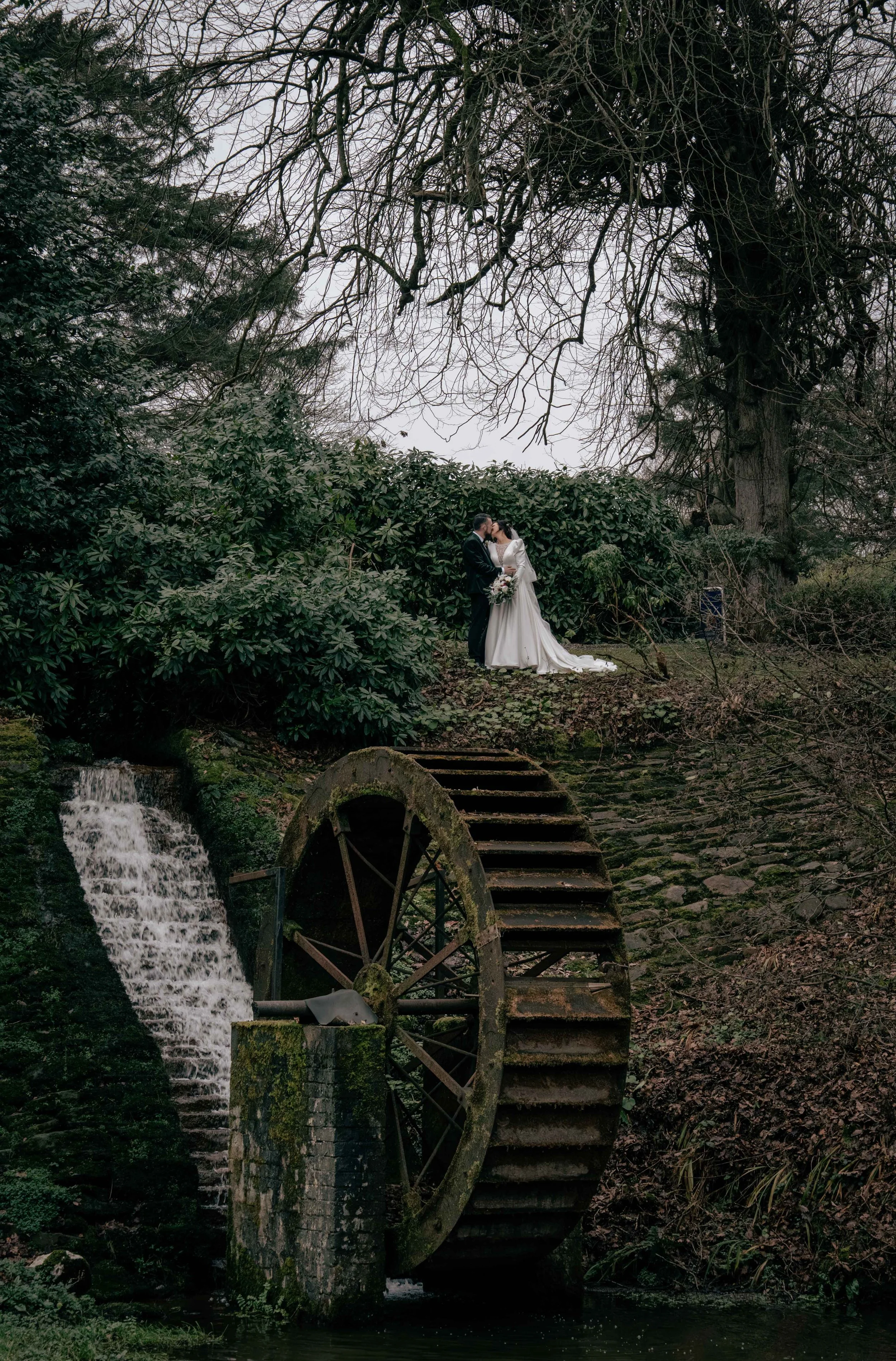 A bride and groom standing kissingly in a lush, green outdoor setting with trees and bushes. They are positioned on a grassy area above an old waterwheel and waterfall, with moss and weathered stones nearby.