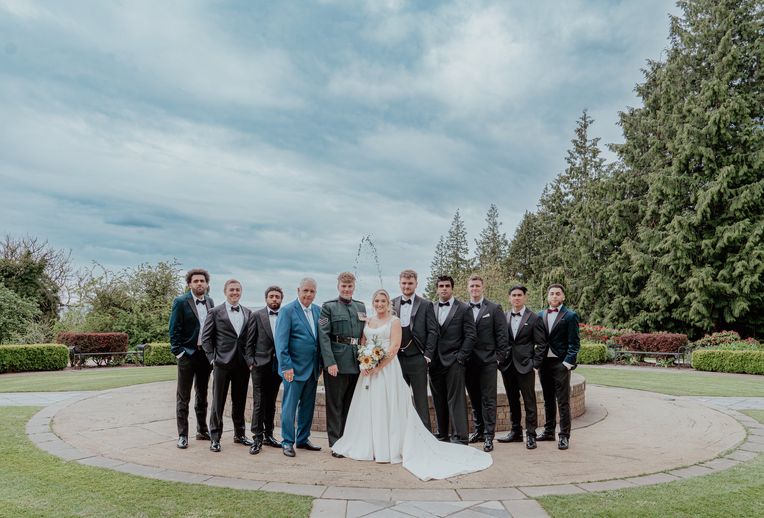 A group photograph of wedding guests and the bride and groom outdoors in a garden, with tall trees and a cloudy sky in the background.