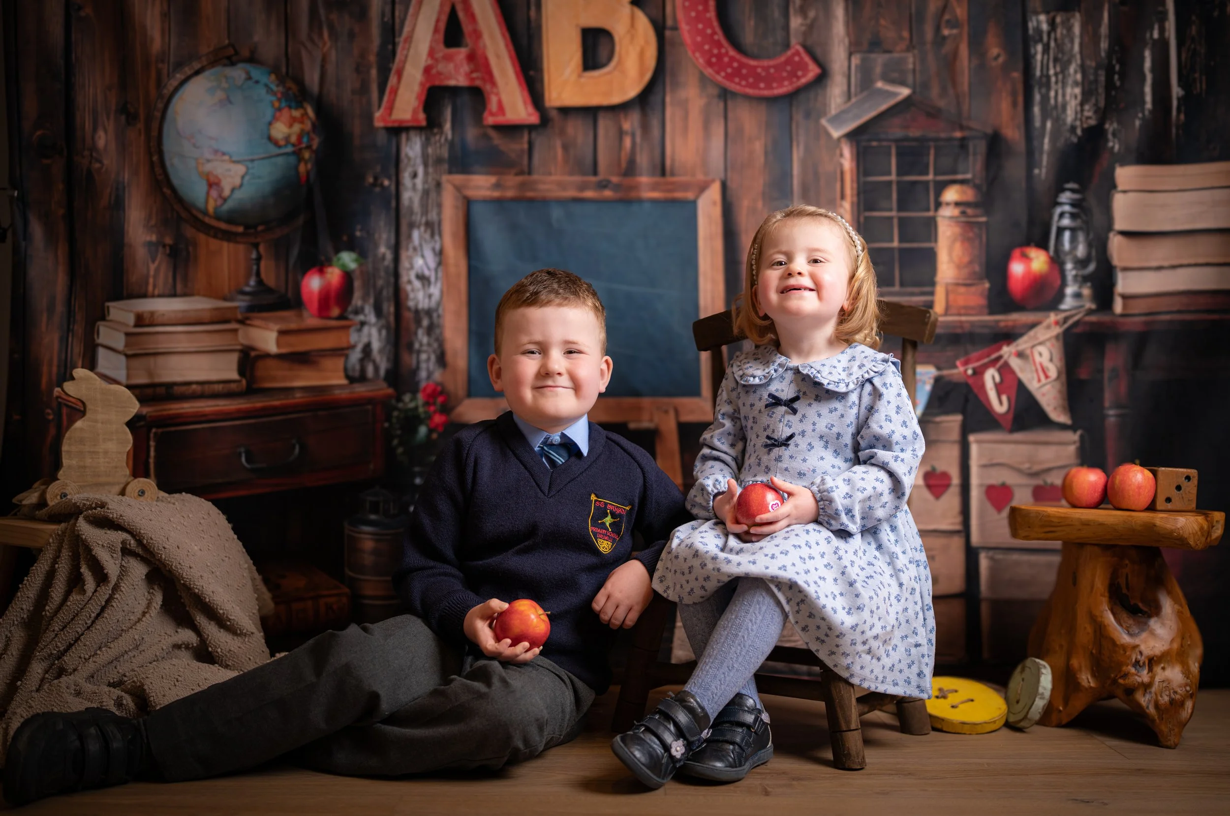 Two young children, a boy and a girl, posing for a photo in a classroom-themed setting. The boy is seated on the floor, wearing a navy sweater with a school crest, and holding an apple. The girl is sitting on a small wooden chair, wearing a light blue dress with a floral pattern, and holding an apple. The background features educational decorations, such as a globe, books, and a chalkboard, with large colorful letters spelling 'ABC' on the wall.