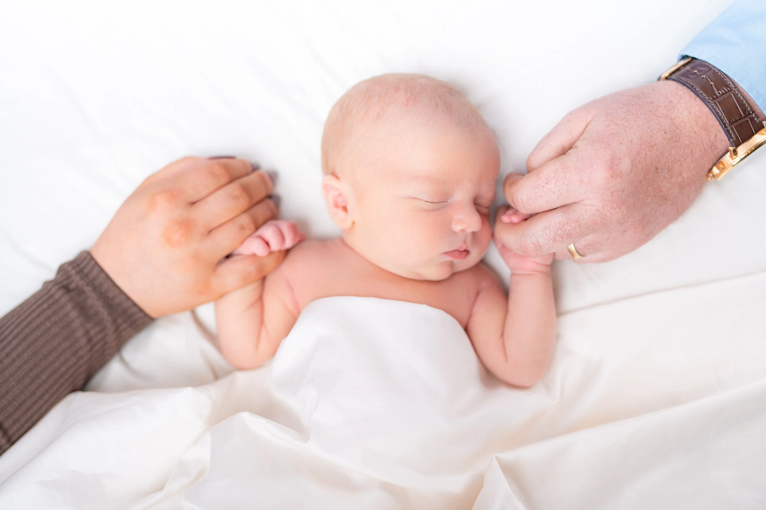 A newborn baby sleeping on a white surface, with hands gently holding the baby's head and hand.