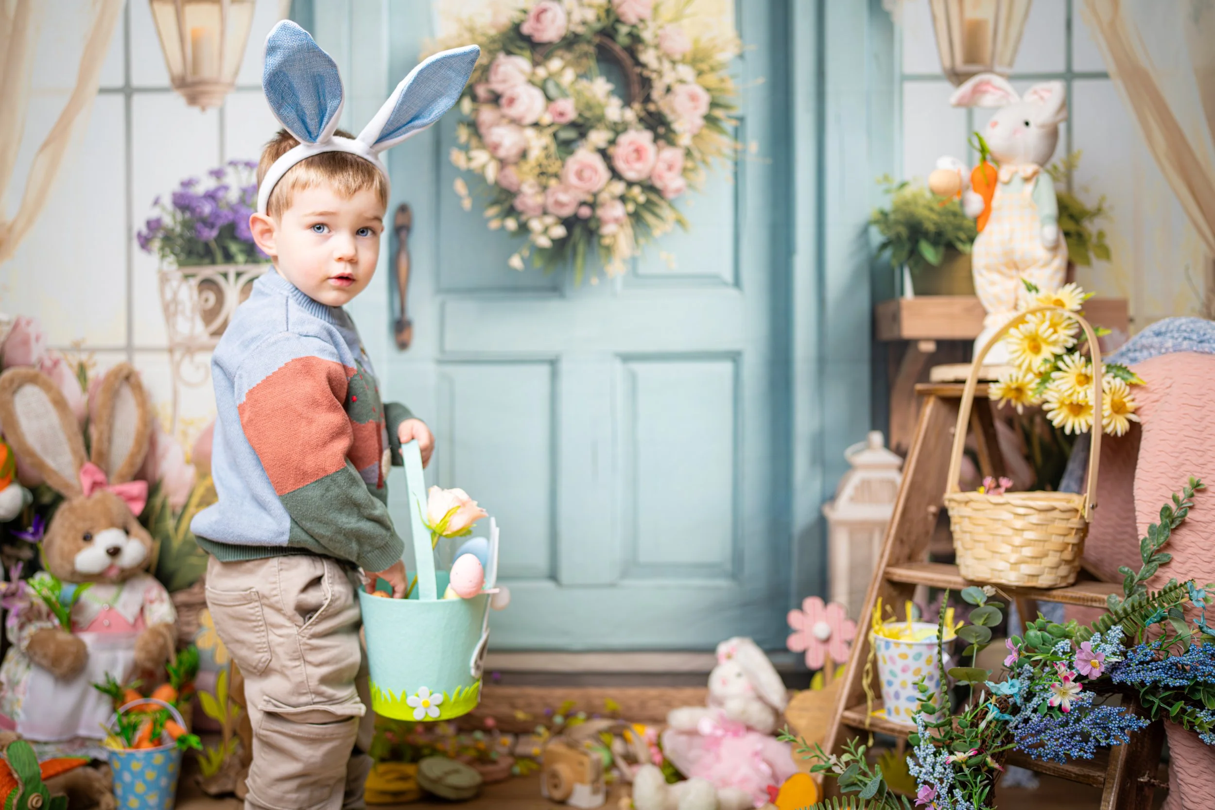Young boy with bunny ears hat, holding an Easter basket filled with eggs and flowers, in a decorated spring-themed setting with plush bunnies, flowers, and Easter decorations.