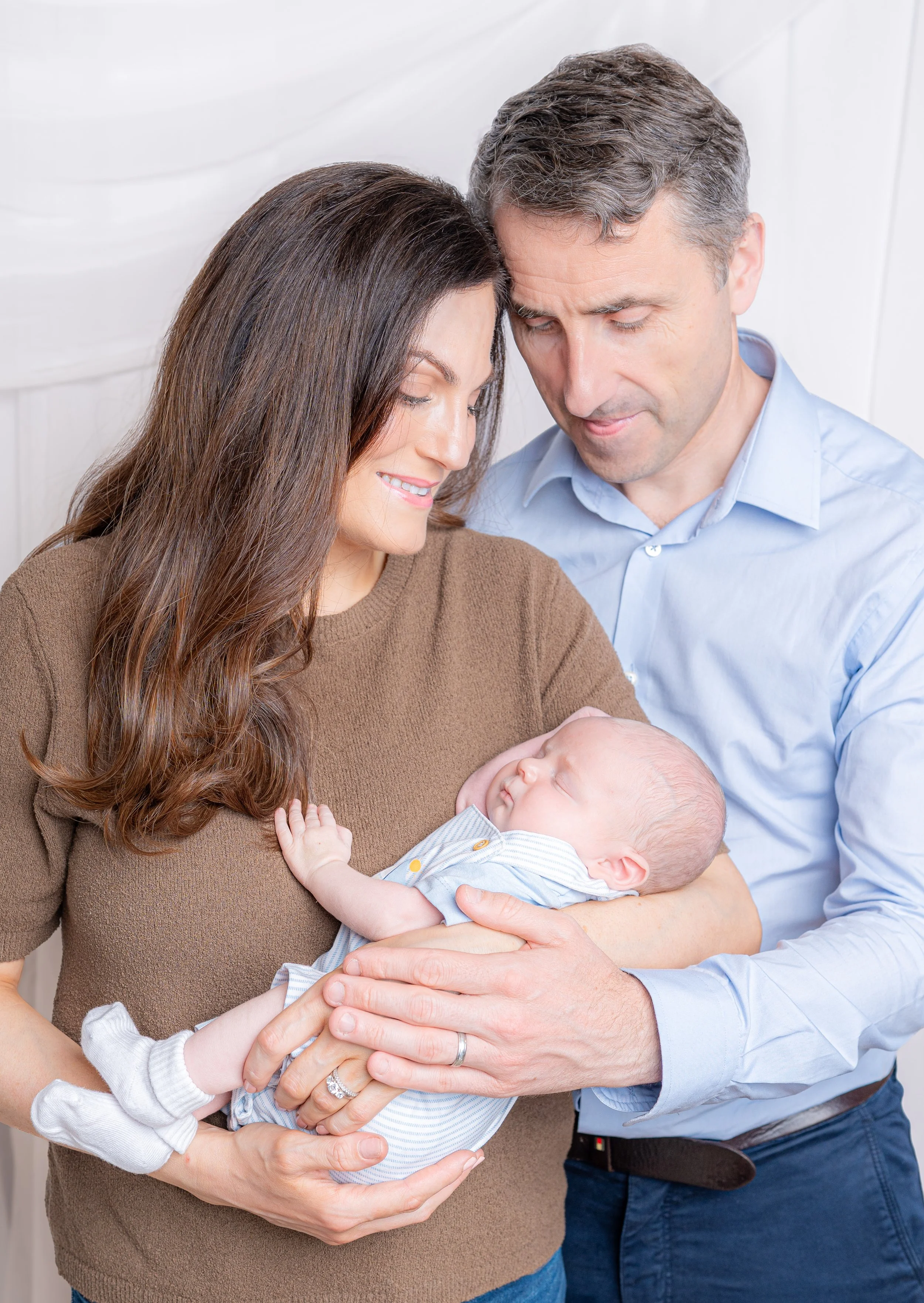 A smiling woman in a brown sweater holding a sleeping baby in a striped outfit, with her hand supporting the baby's head and hand. A man with gray hair in a light blue shirt gently holds the baby's bottom, both leaning in close with foreheads touching in a tender moment.