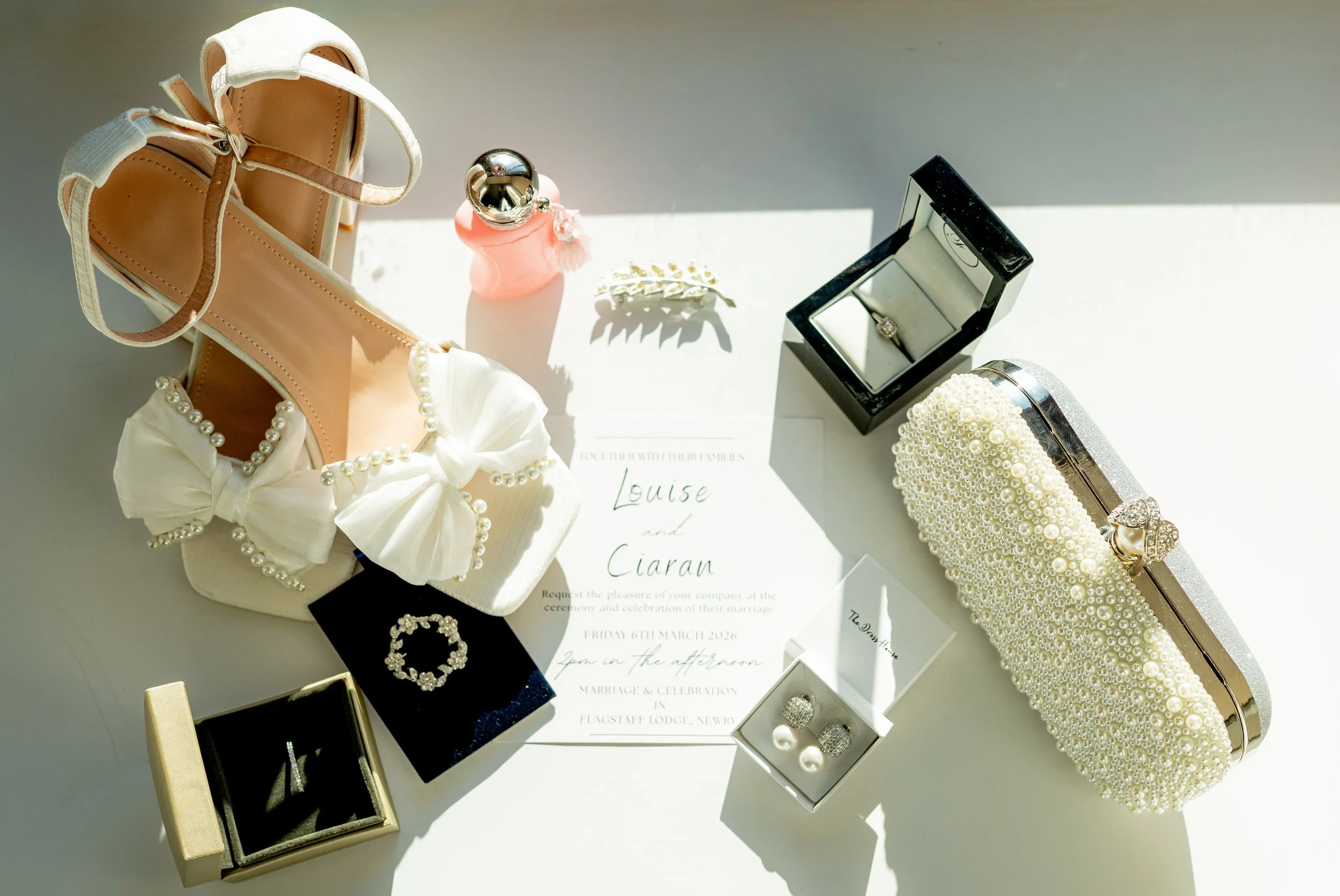 Wedding invitation surrounded by pearl jewelry, a high-heeled shoe, pearl-embellished handbags, and jewelry boxes with rings and earrings on a white surface.