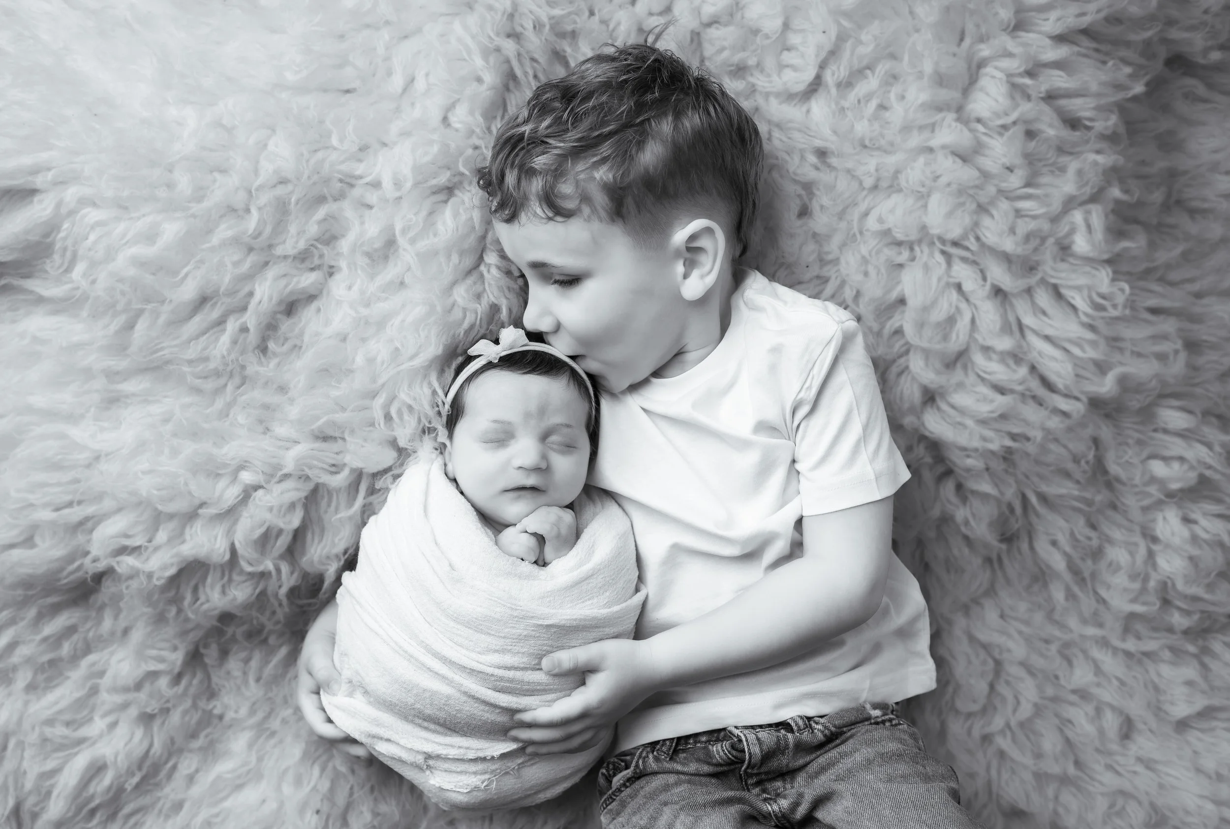 A young boy with curly hair holding a sleeping baby girl wrapped in a blanket, lying on a fluffy surface.