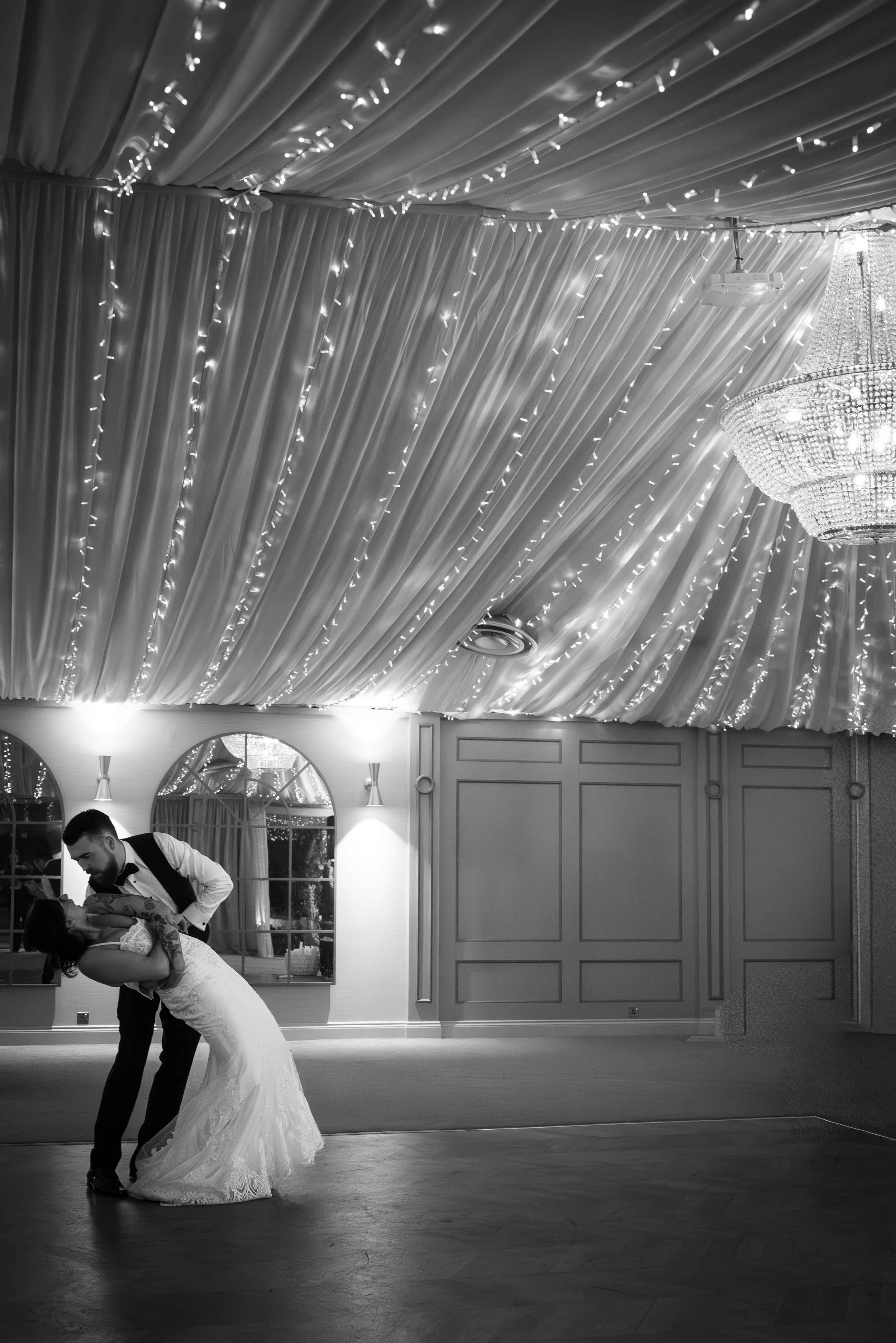 A bride and groom sharing a dance in a decorated wedding reception hall with draped ceiling, string lights, and a large chandelier.