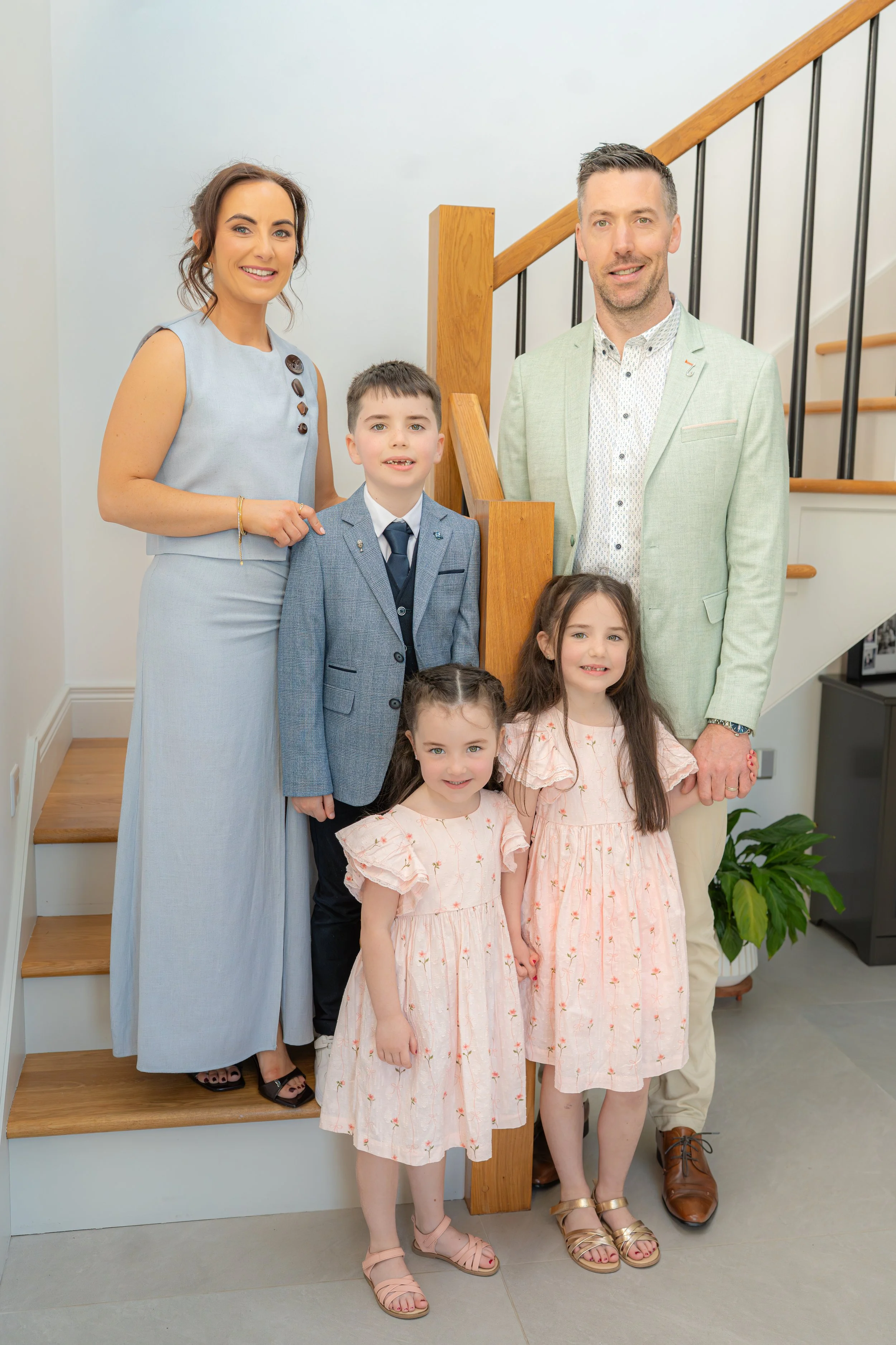 A group of six people, two adults and four children, standing on a staircase interior with a white background. The woman on the left is wearing a light blue dress with dark buttons and black shoes, and the man on the right is wearing a light green blazer with a white patterned shirt and brown shoes. The children are dressed in light pink dresses, gray suit, and casual outfits, smiling and looking at the camera.