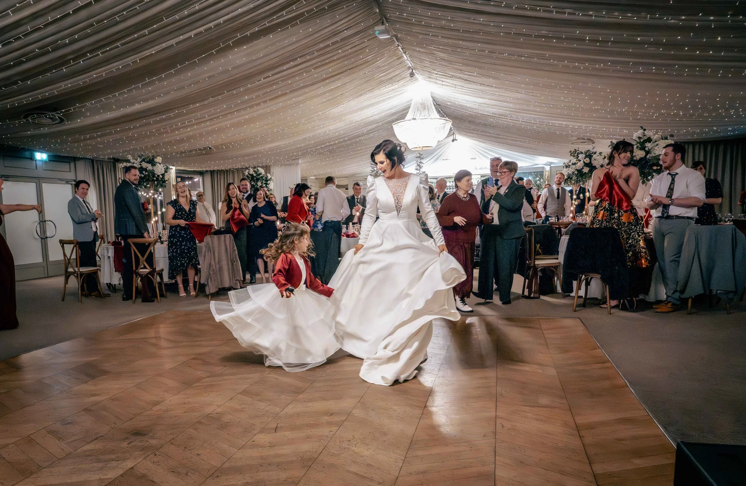 Bride dancing with a young girl in a ballroom decorated with string lights and floral arrangements with guests watching and enjoying the celebration.
