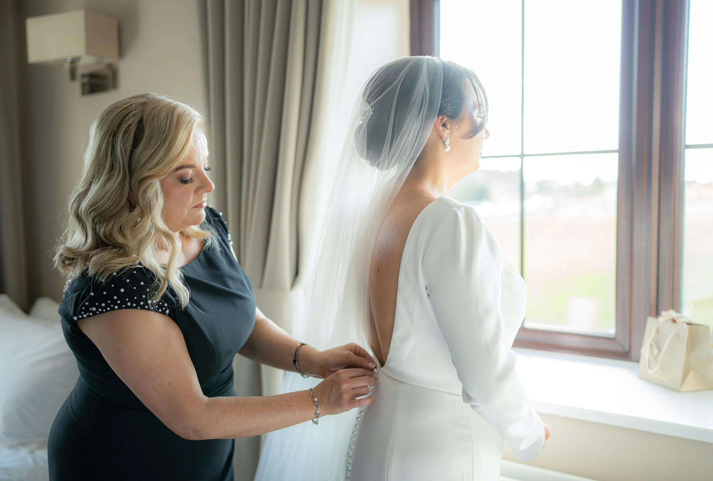 A bride in a white dress with a veil stands by a window, while another woman fastens the back of her dress.