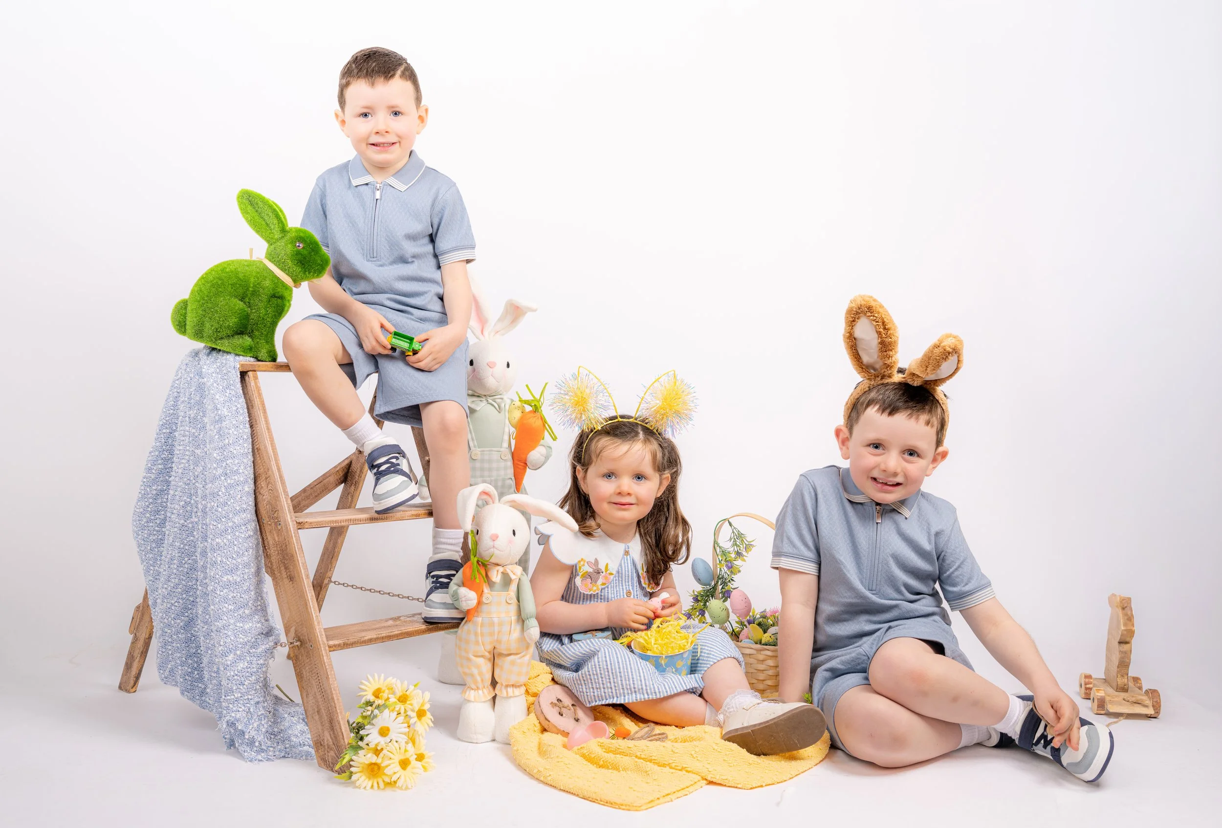 Three children dressed with bunny ears and Easter accessories surrounded by Easter decorations, stuffed bunnies, and flowers