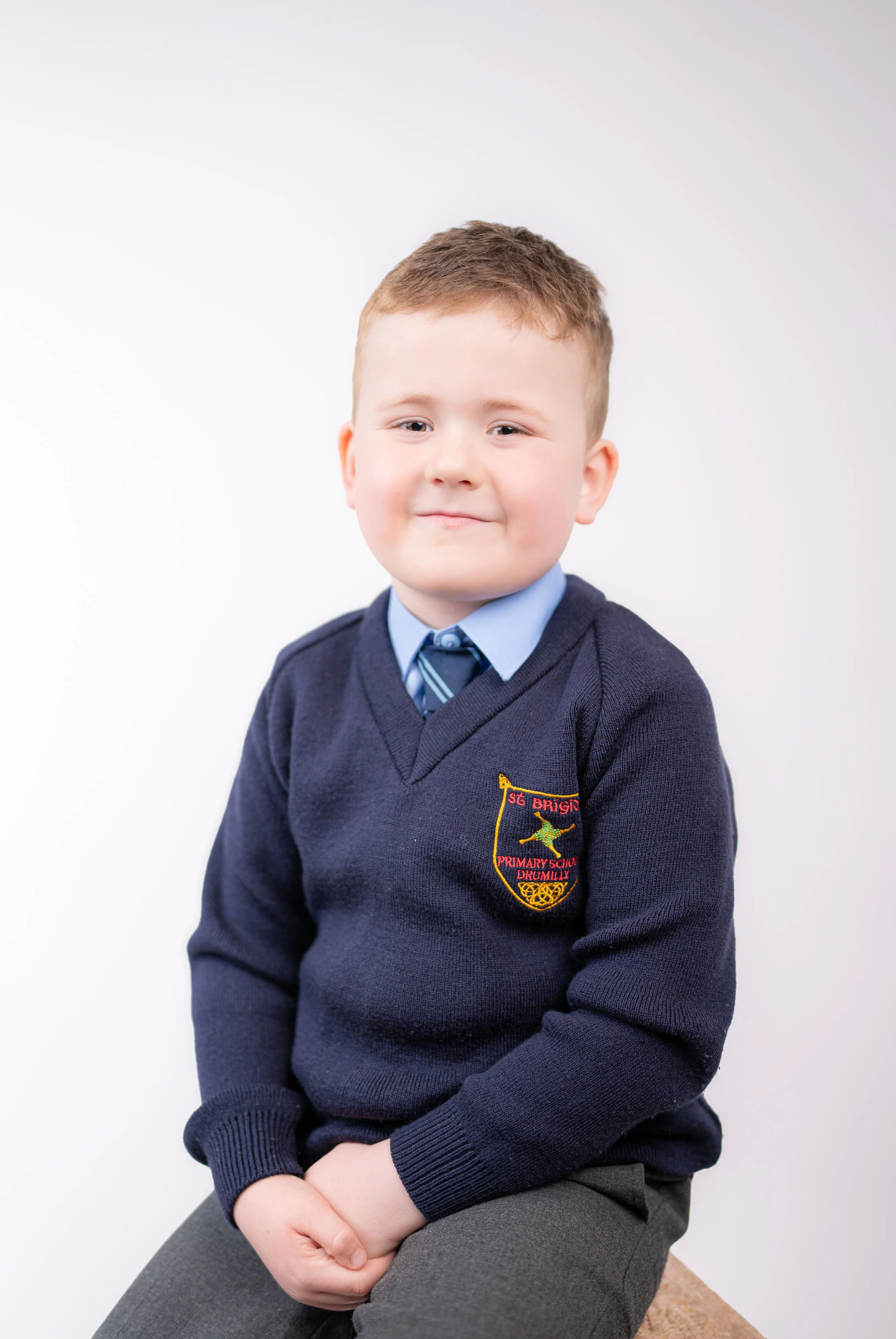 A young boy dressed in a primary school uniform with a navy sweater featuring an emblem from 'SG Brigid Primary School Drumilly', a light blue shirt, and a navy striped tie, sitting against a plain white background.