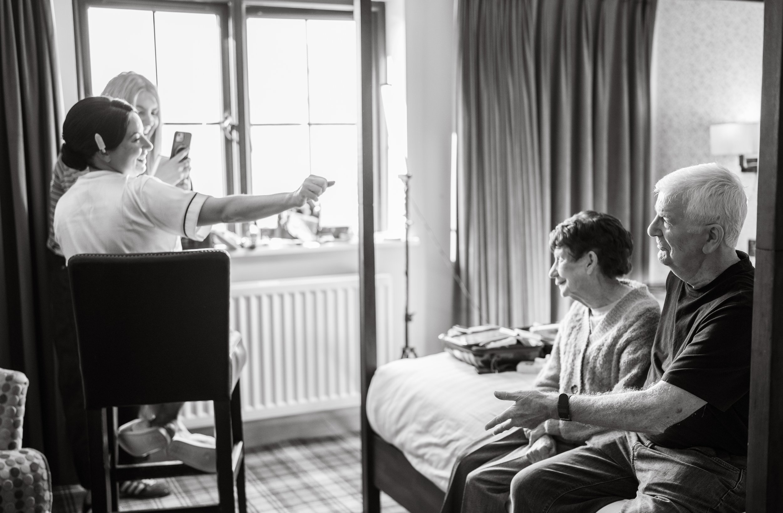 Three women, two elderly women and one younger woman, sitting in a room with large window and curtains. The younger woman, dressed like a nurse or caregiver, is taking a photo or video of the elderly couple with her smartphone. The elderly couple is engaged in conversation, sitting on a bed with stacked papers or books nearby.